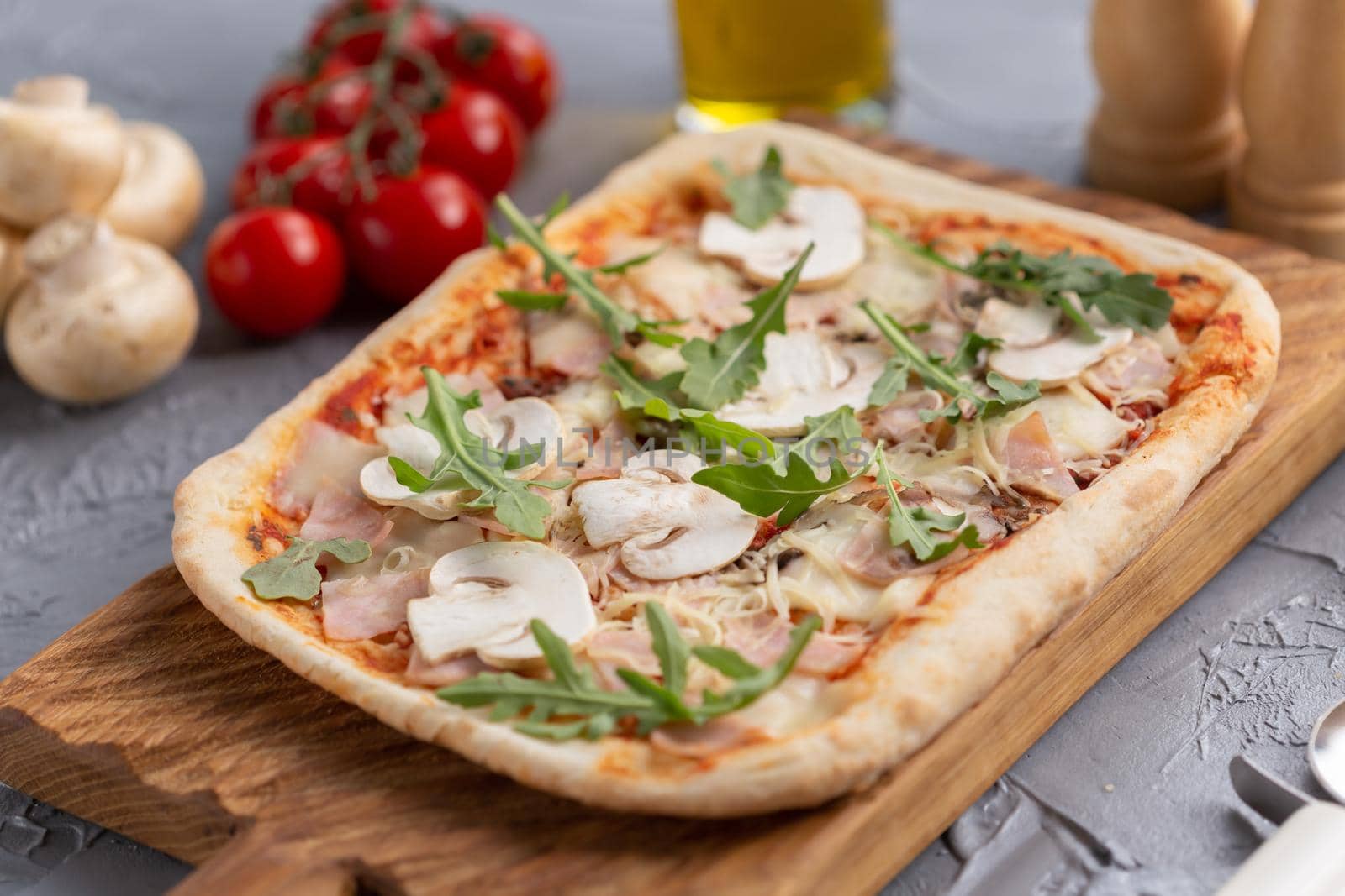 Rectangular pizza with mushrooms, tomatoes and arugula on a wooden cutting board.