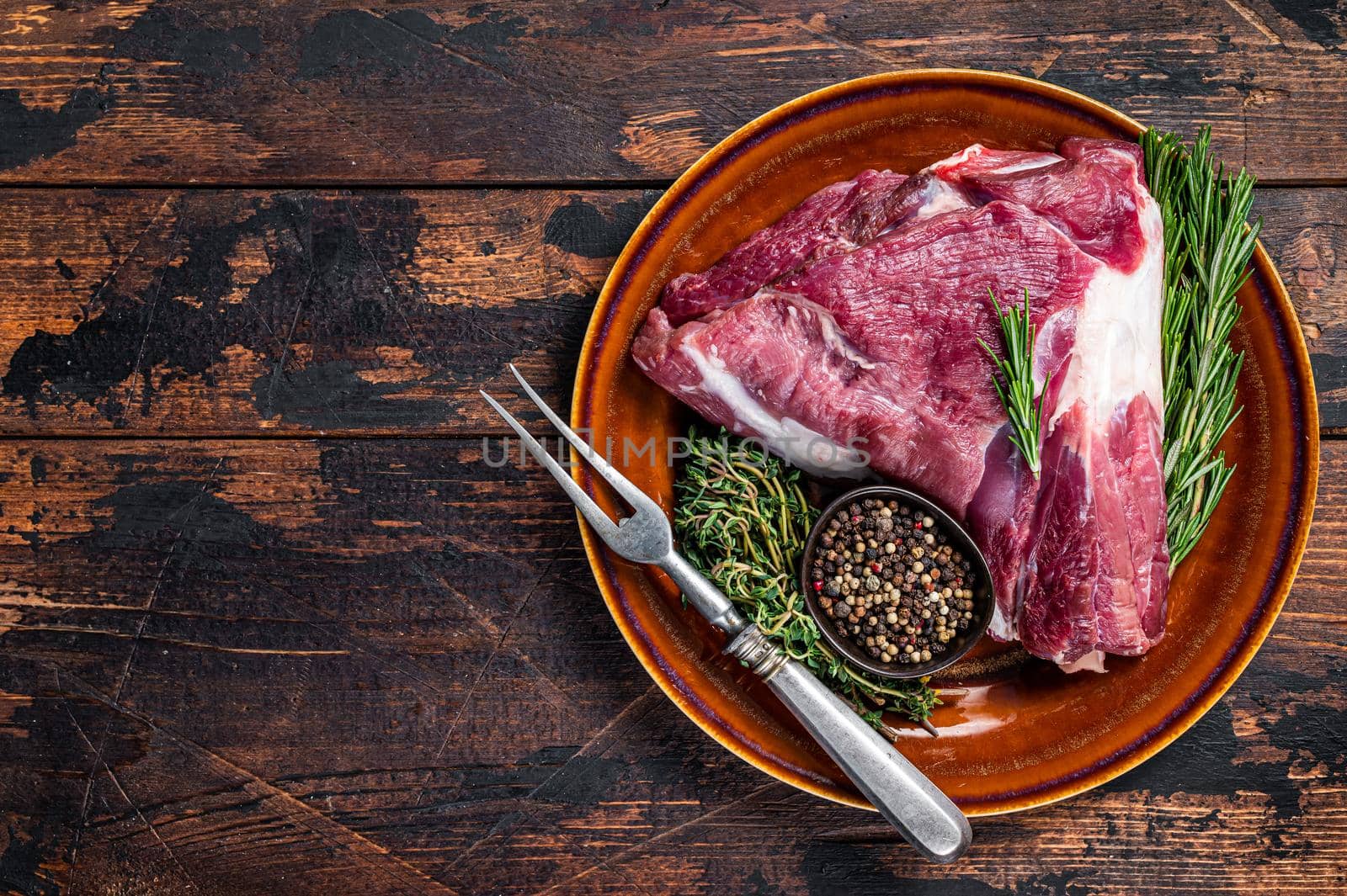 Raw lamb mutton thigh or leg with rosemary and thyme in rustic plate. Dark wooden background. Top view. Copy space.