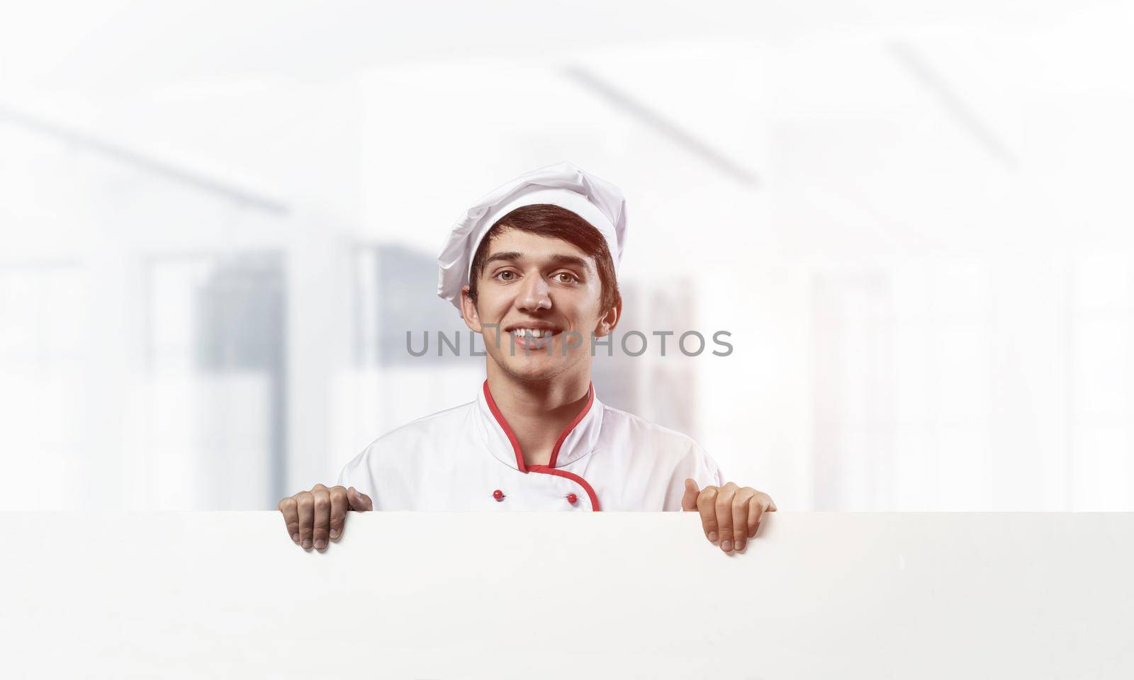 Young male chef with blank white signboard. Handsome chef in white and red uniform on light room blurred background. Cooking classes announcement. Professional catering service concept with copy space