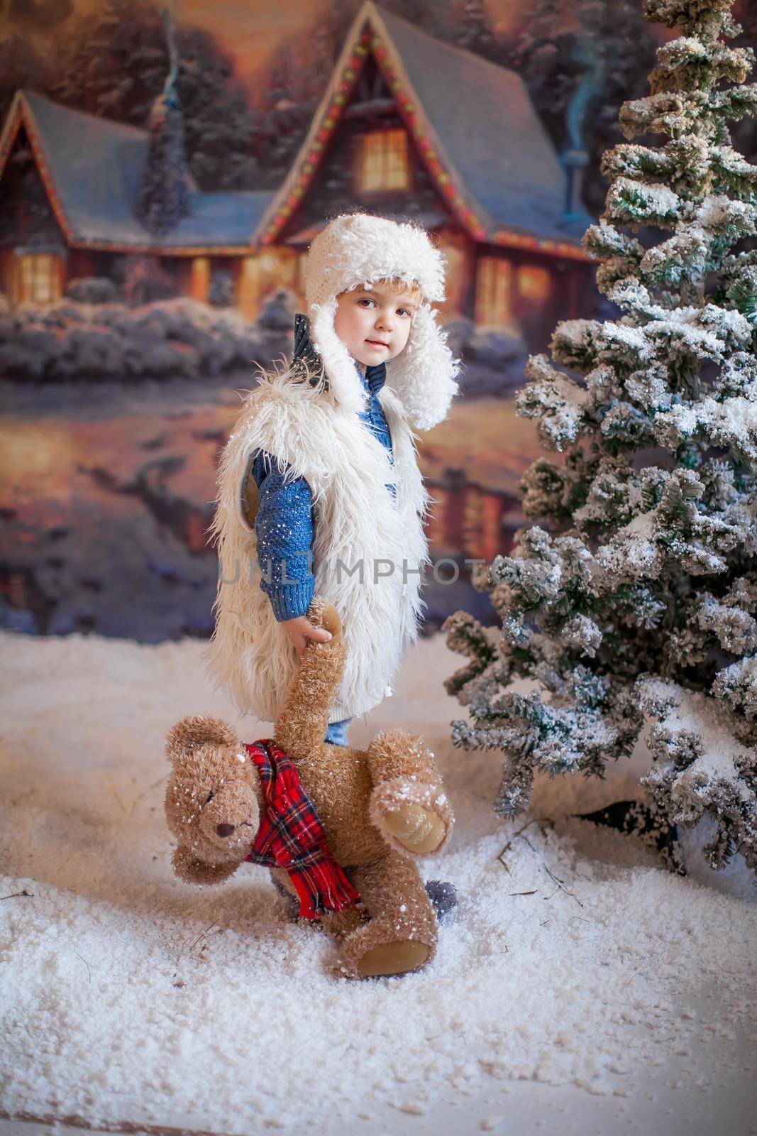 A boy in a white hat and boots on the background of an artificial forest in the winter village
