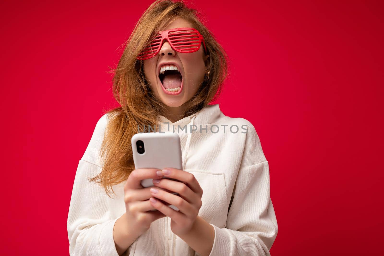 Beautiful emotional young blonde woman wearing stylish white hoodie and funny colorful glasses standing isolated over colourful background holding and using mobile phone communicating online on the internet and yawning.