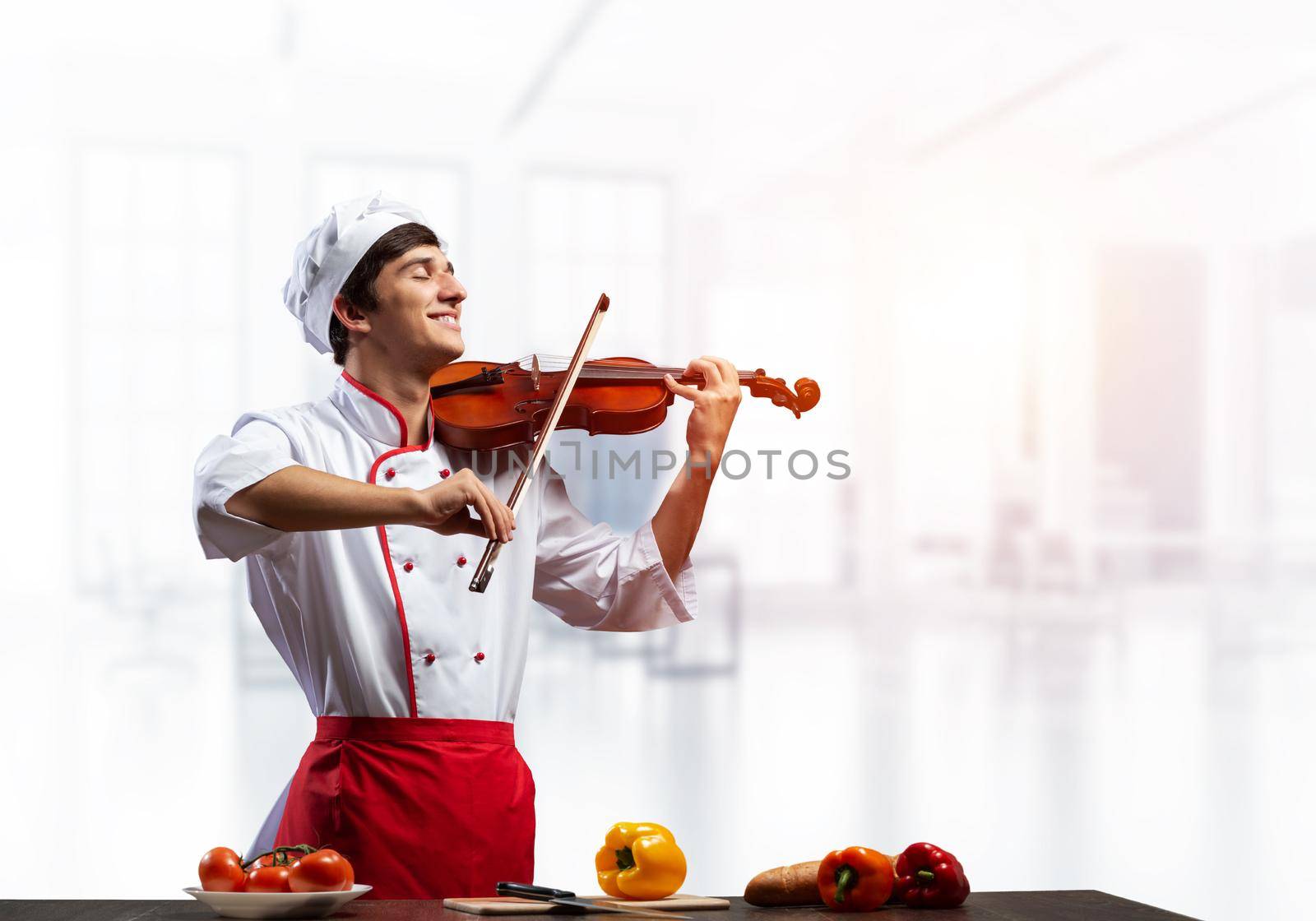 Young male chef with violin standing near cooking table with vegetables. Handsome chef in white hat and red apron in light kitchen interior. Creative culinary performance. High cooking skills concept