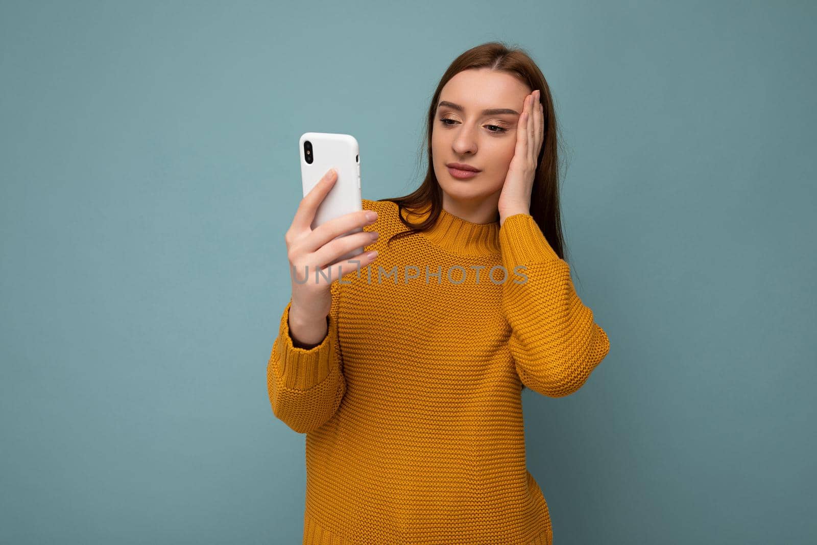 Photo of beautiful surprised young woman wearing orange sweater poising isolated on blue background with empty space holding in hand and using mobile phone communicating online with friends looking at gadjet screen.