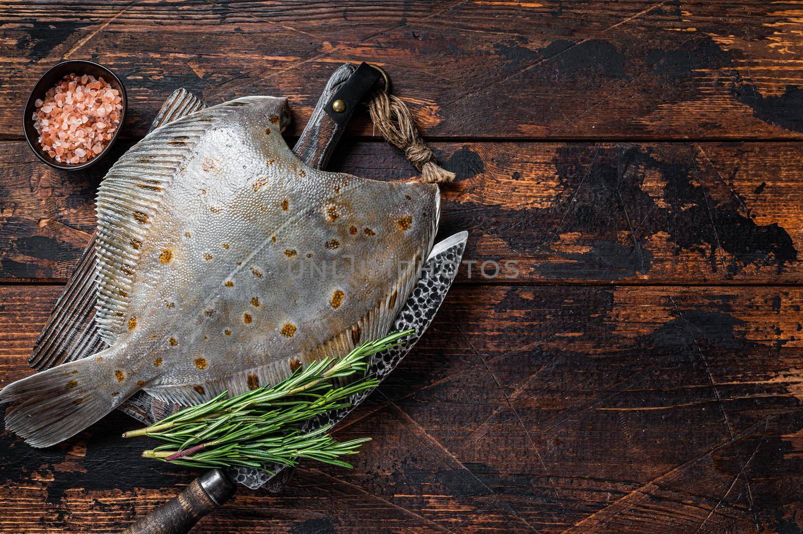 Raw plaice flatfish fish on butcher board with knife. Dark wooden background. Top view. Copy space.