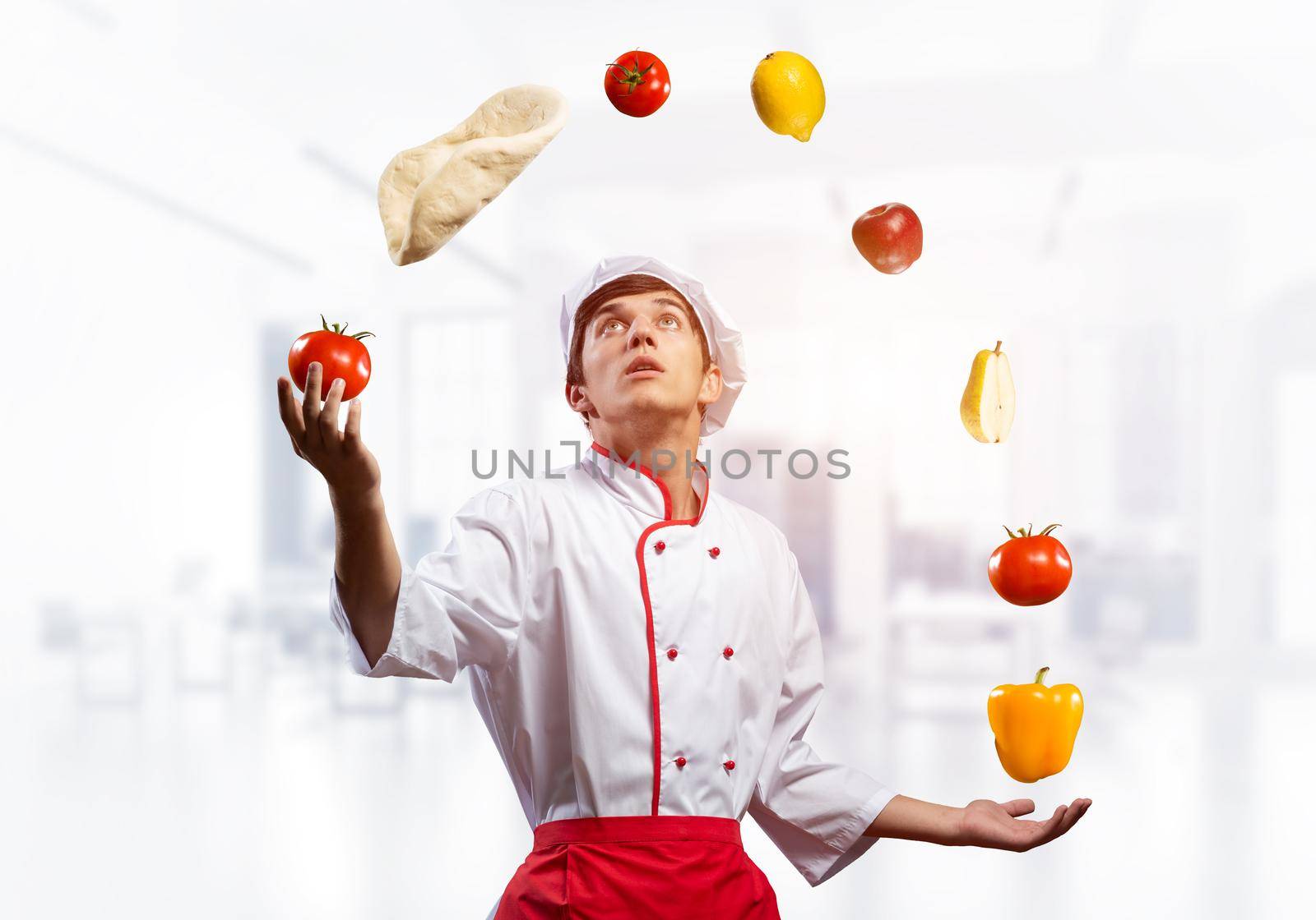 Young male chef juggles with food ingredients. Handsome chef in white hat and red apron in light kitchen interior. Professional cooking classes advertising. Food retail campaign announcement.