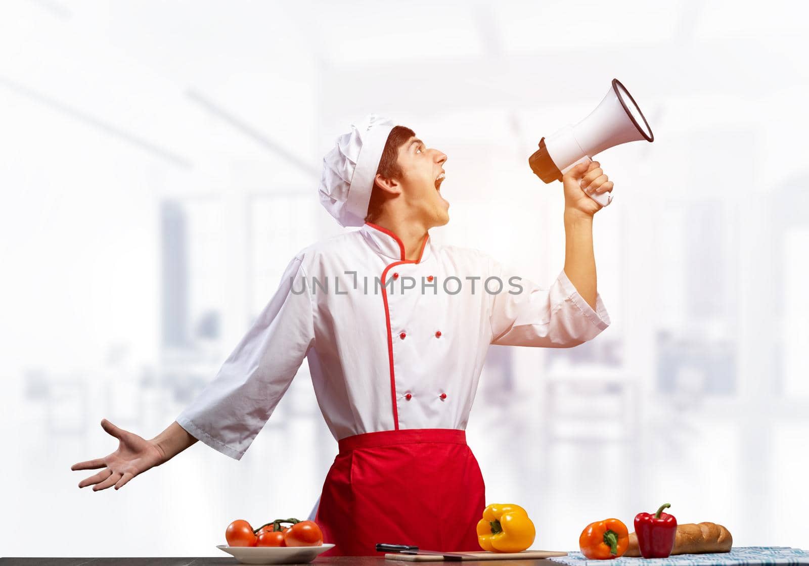 Young chef shouting loudly into megaphone. Emotional caucasian chef in white hat and red apron on light blurred background. Restaurant advertisement, food retail campaign announcement.