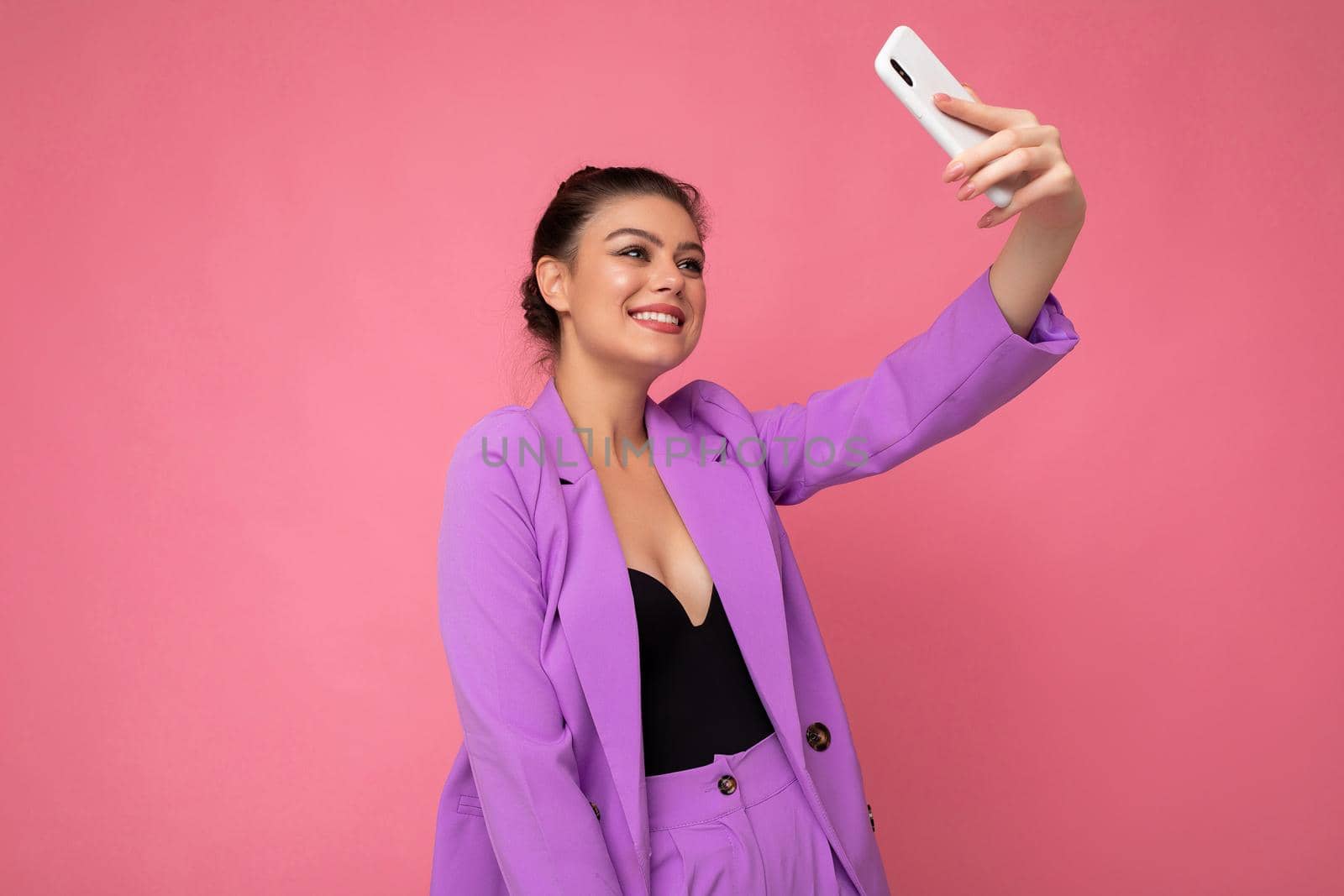Photo of pretty young woman wearing purple suit taking selfie photo on the mobile phone isolated over pink background looking at phone camera.Copy space