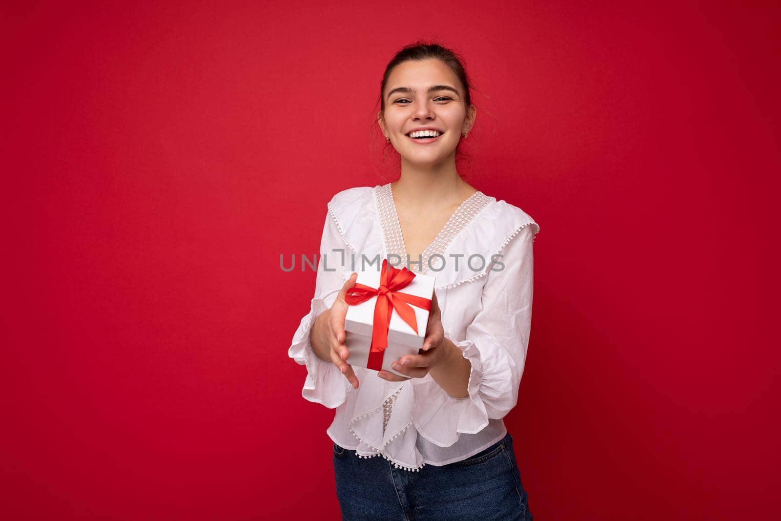 Beautiful happy young brunette woman isolated over colourful background wall wearing stylish casual clothes holding gift box and looking at camera. Empty space