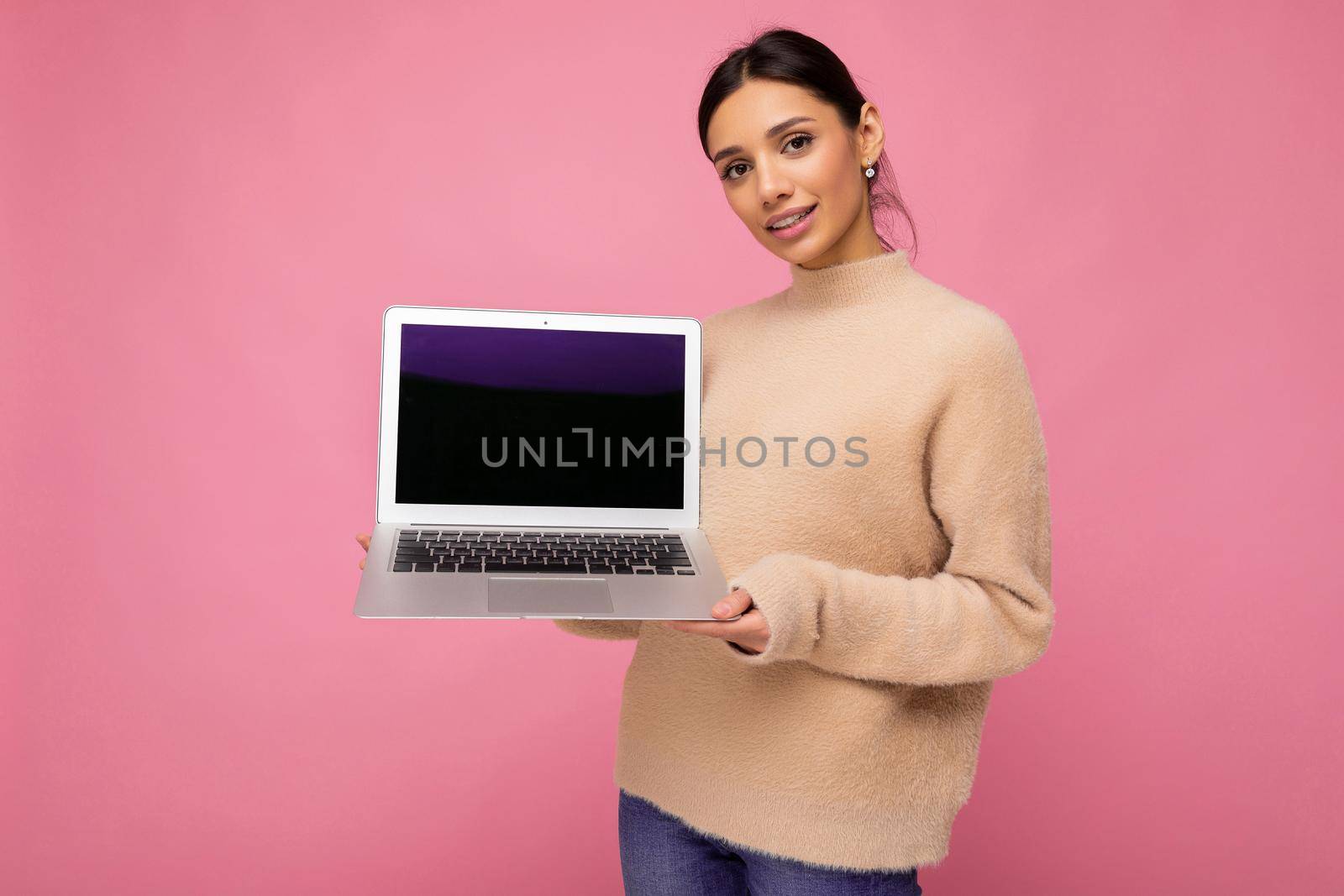 beautiful smiling young woman with dark hair looking at camera holding computer laptop with empty monitor screen with mock up and copy space wearing light colour sweater isolated on pink wall background.