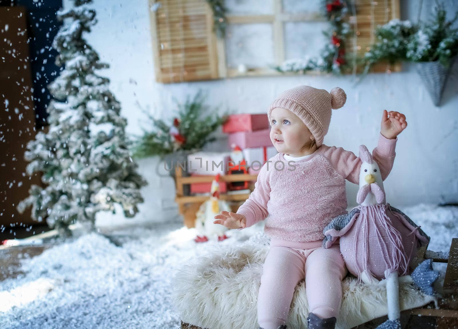 A baby girl sits in a Studio with artificial winter scenery in a pink sweater and a pink beanie by ElenaBatkova