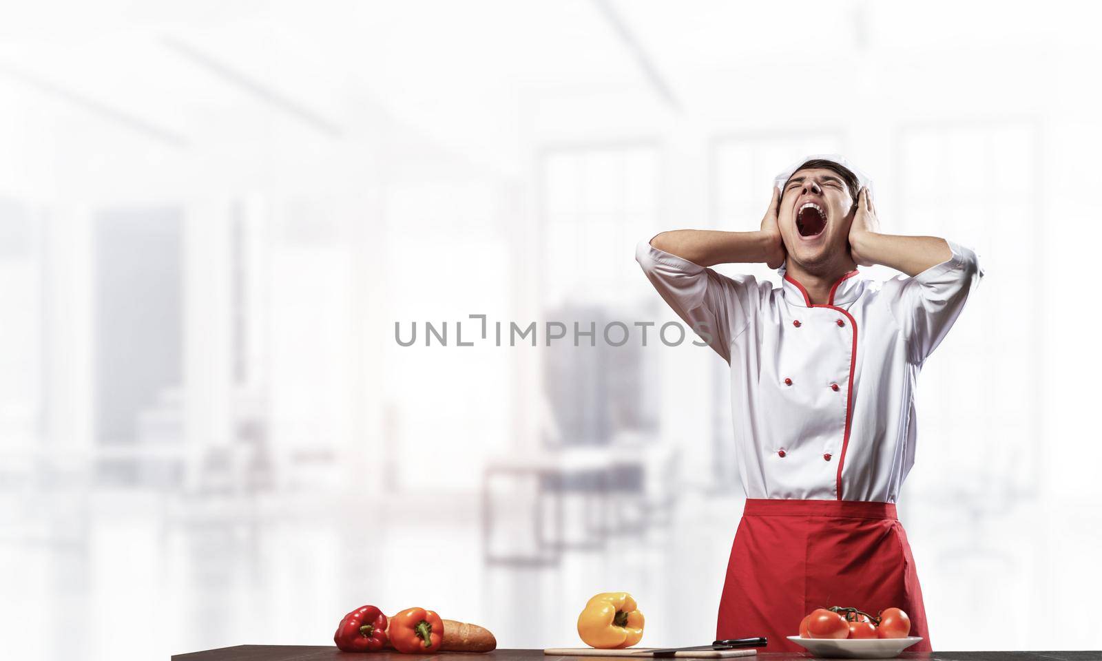 Young male chef screaming in shock and horror. Emotional caucasian chef in white hat and red apron standing with open mouth in light kitchen interior. Creative marketing campaign, big discounts.