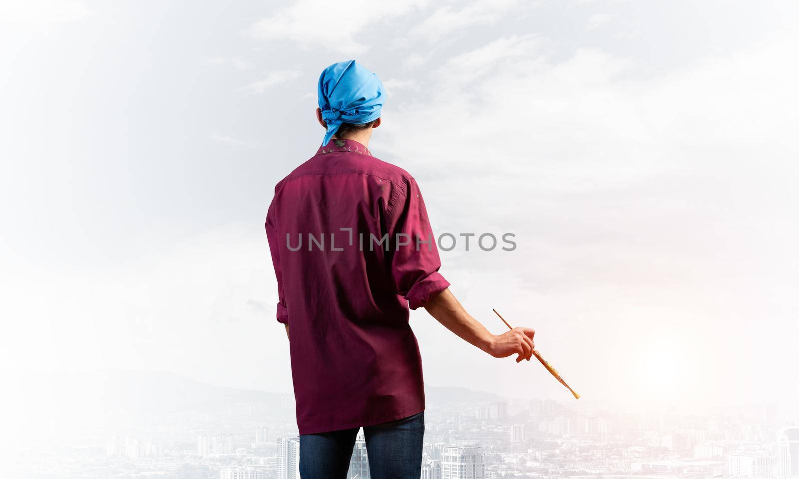 Young male painter artist holding paintbrush. Back view of painter in shirt and bandana on background cloudy sky and cityscape. Creative hobby and artistic occupation. Art school courses concept.