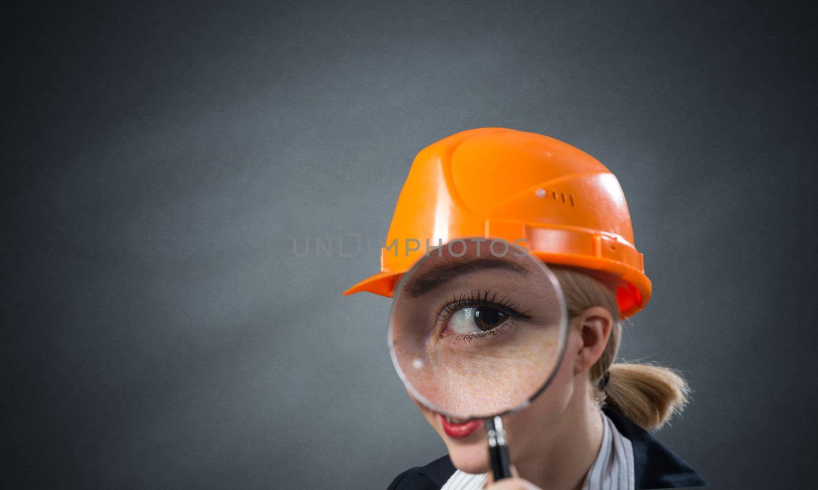 Technical inspector looking through magnifying glass. Woman civil engineer in orange safety helmet checking with magnifier on grey wall background. Commercial real estate appraisal and consultation.
