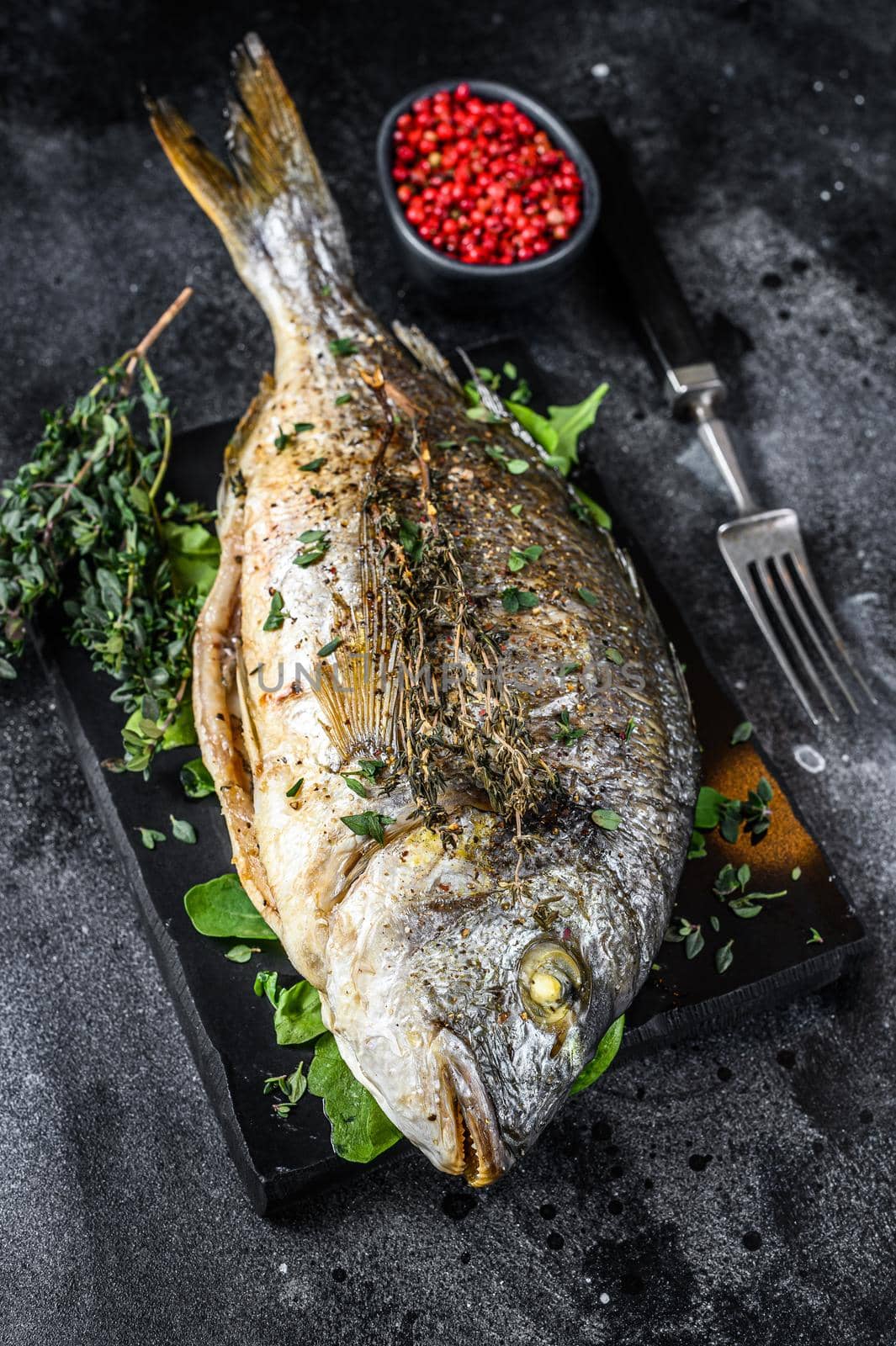 Roasted sea bream fish with herbs on a cutting board. Black background. Top view.