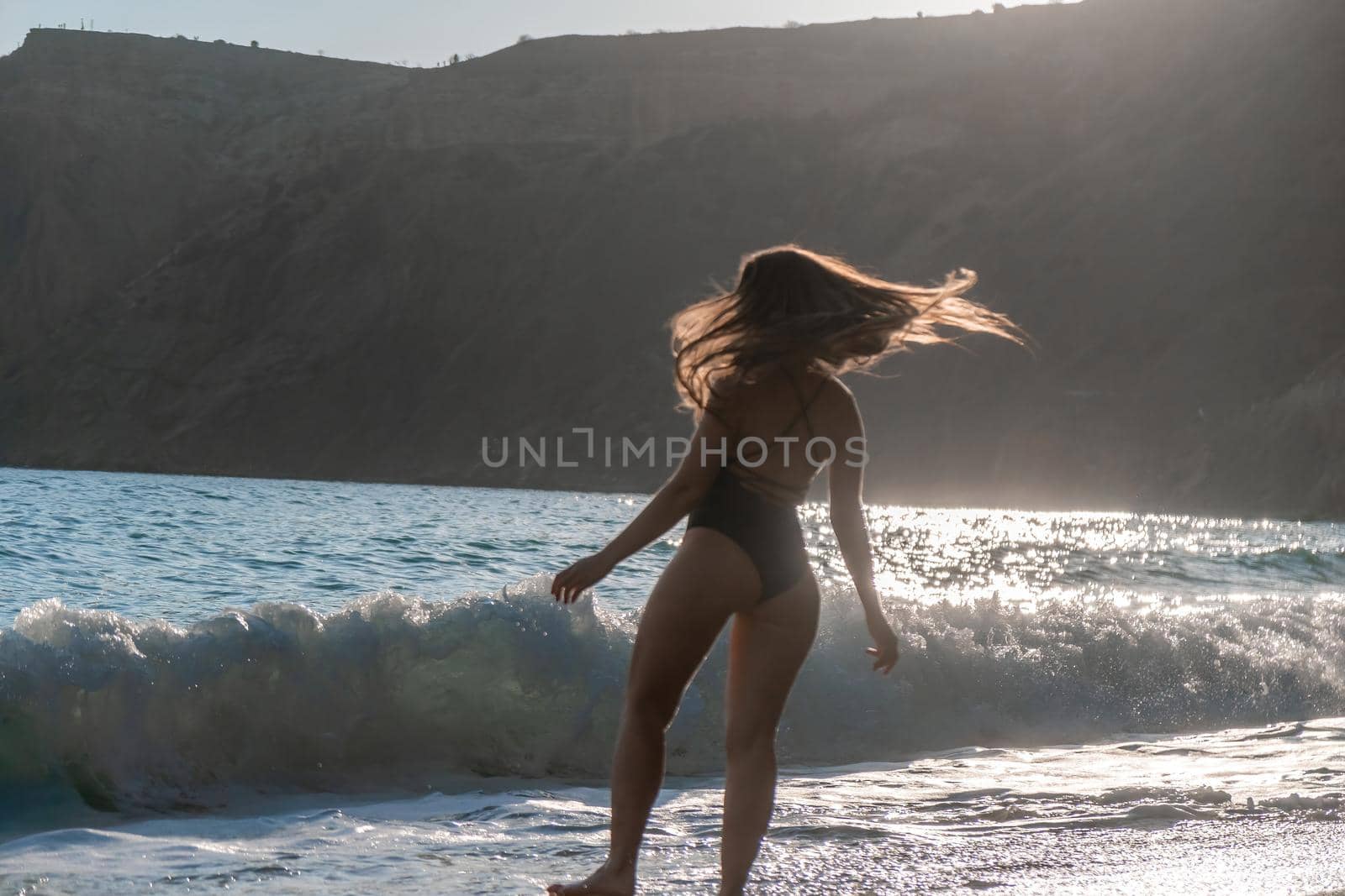 Selective focus. Happy carefree sensual woman with long hair in black swimwear posing at sunset beach. Silhouette of young beautiful playful positive woman outdoor. Summer vacation and trip concept.