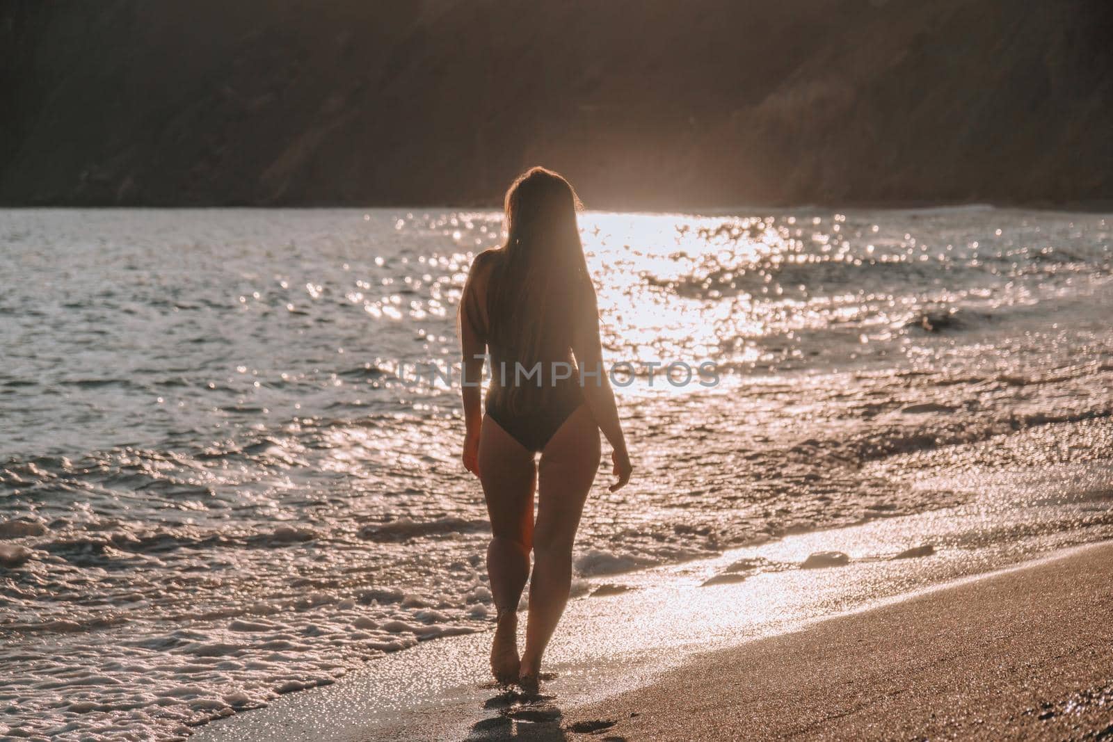 Selective focus. Happy carefree sensual woman with long hair in black swimwear posing at sunset beach. Silhouette of young beautiful playful positive woman outdoor. Summer vacation and trip concept.