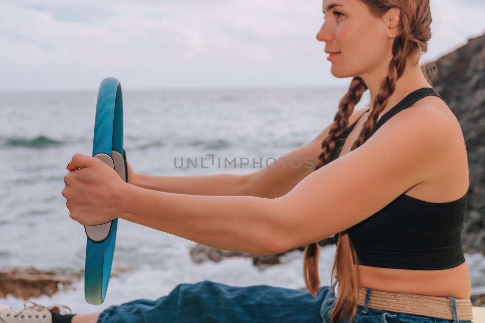 Young woman with long hair, fitness instructor in black Sportswear Leggings and Tops, stretching on a yoga mat with magic pilates ring near the sea on a sunny day, female fitness yoga routine concept.