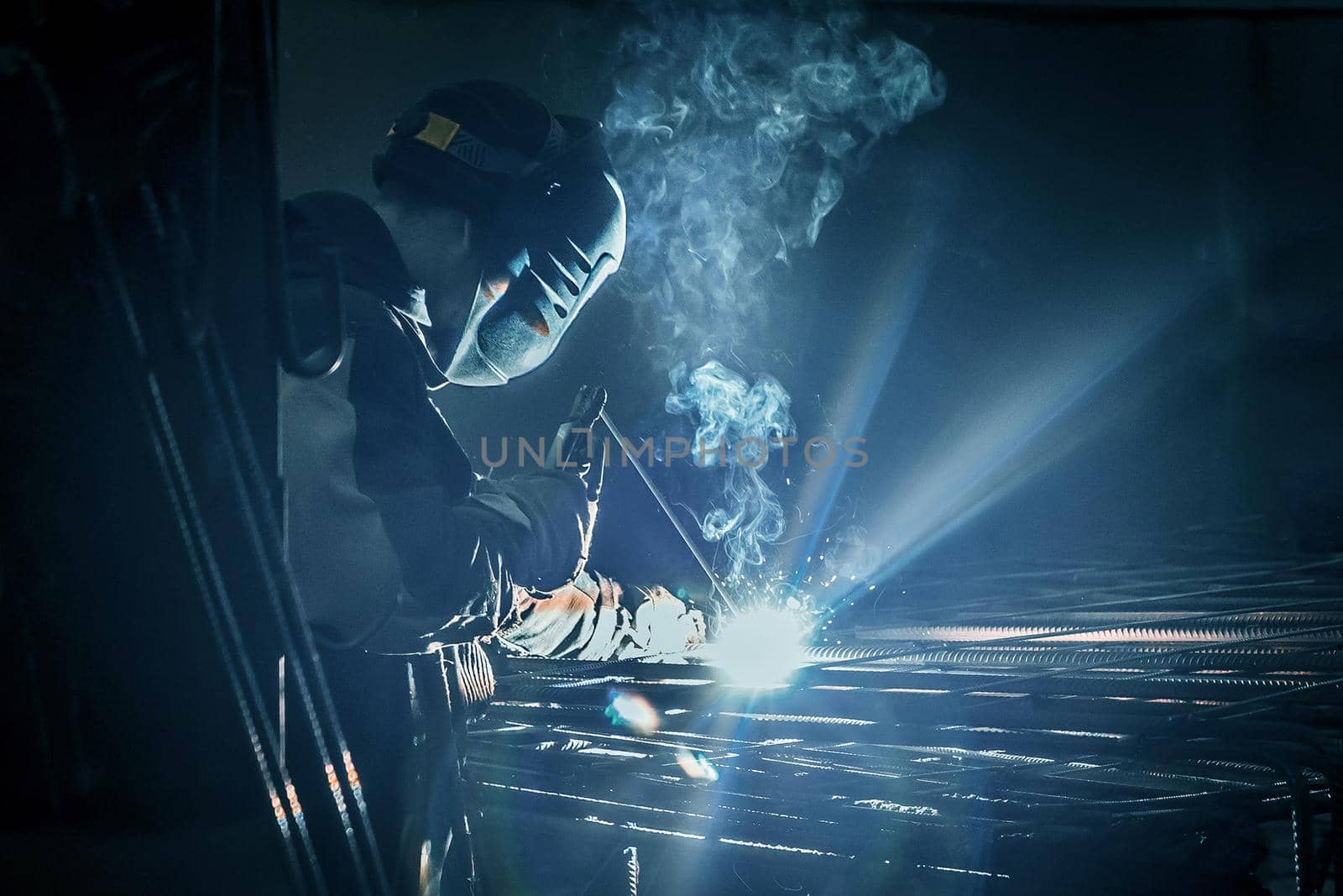 A masked working man is doing welding work on metal structures in a factory or industrial enterprise.