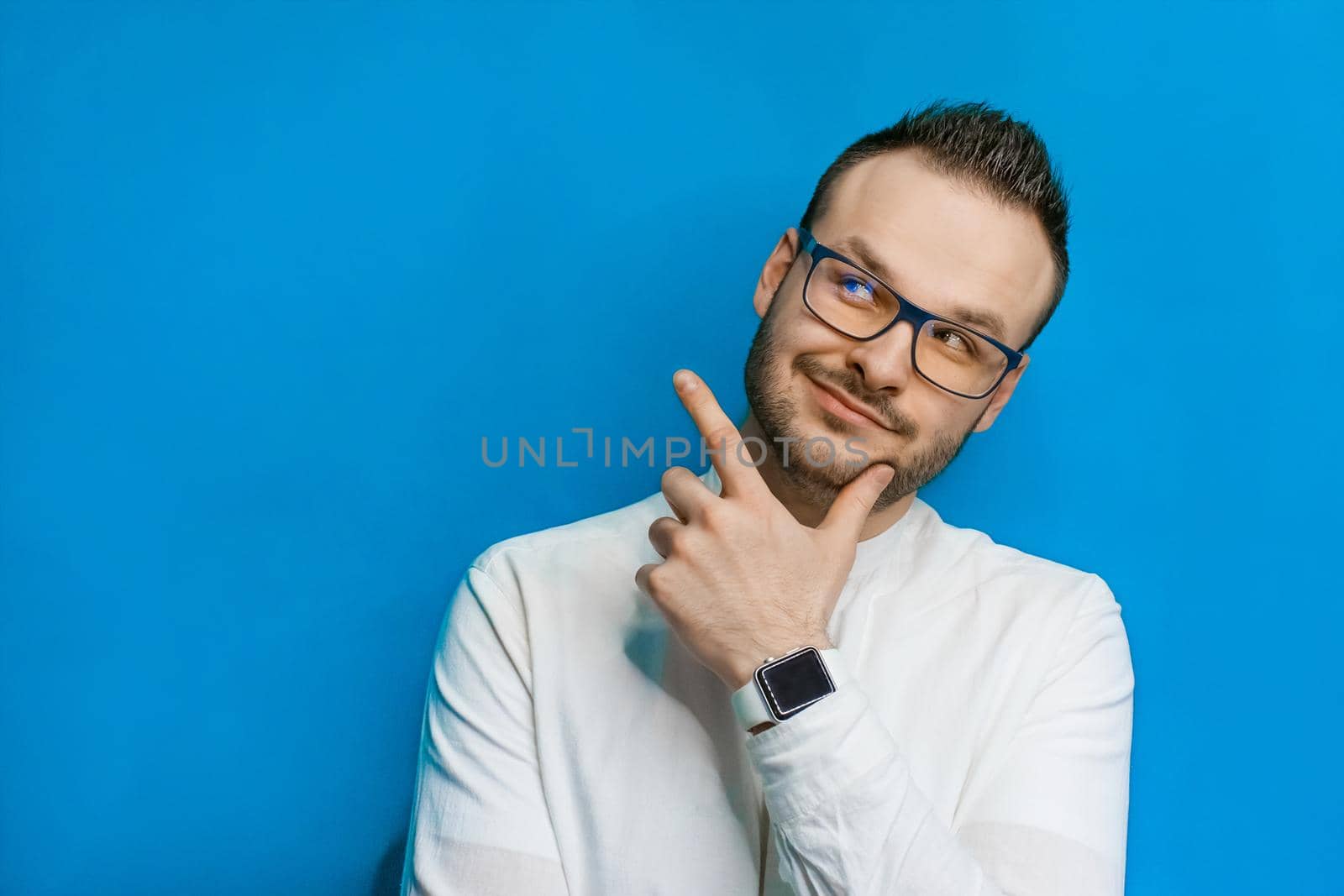 Portrait of a young attractive european businessman in a white shirt, glasses, with a beard and smart watch on a blue background.