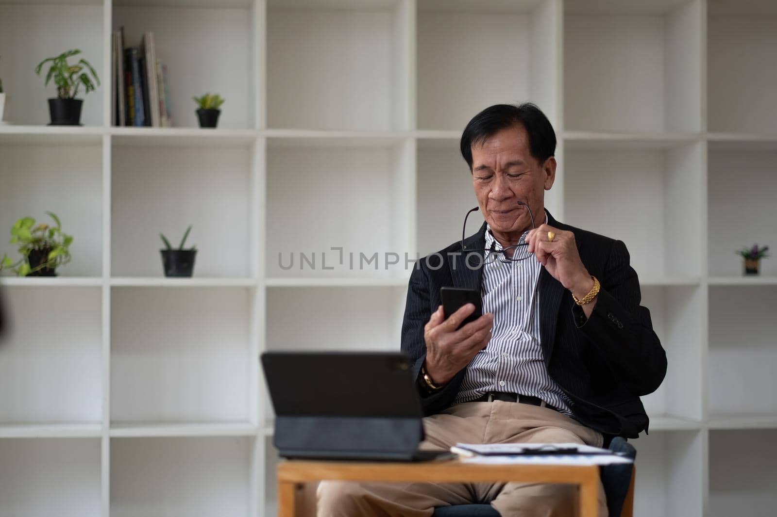 Asian old elder senior man on mobile smart phone and digital tablet on wooden desk.