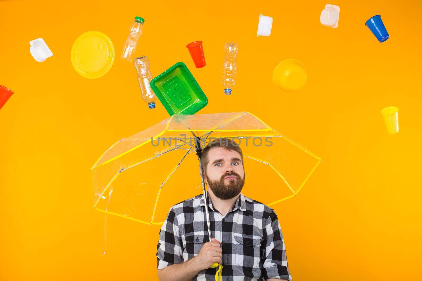 Problem of trash, plastic recycling, pollution and environmental concept - Sad man is standing under trash with umbrella on yellow background.