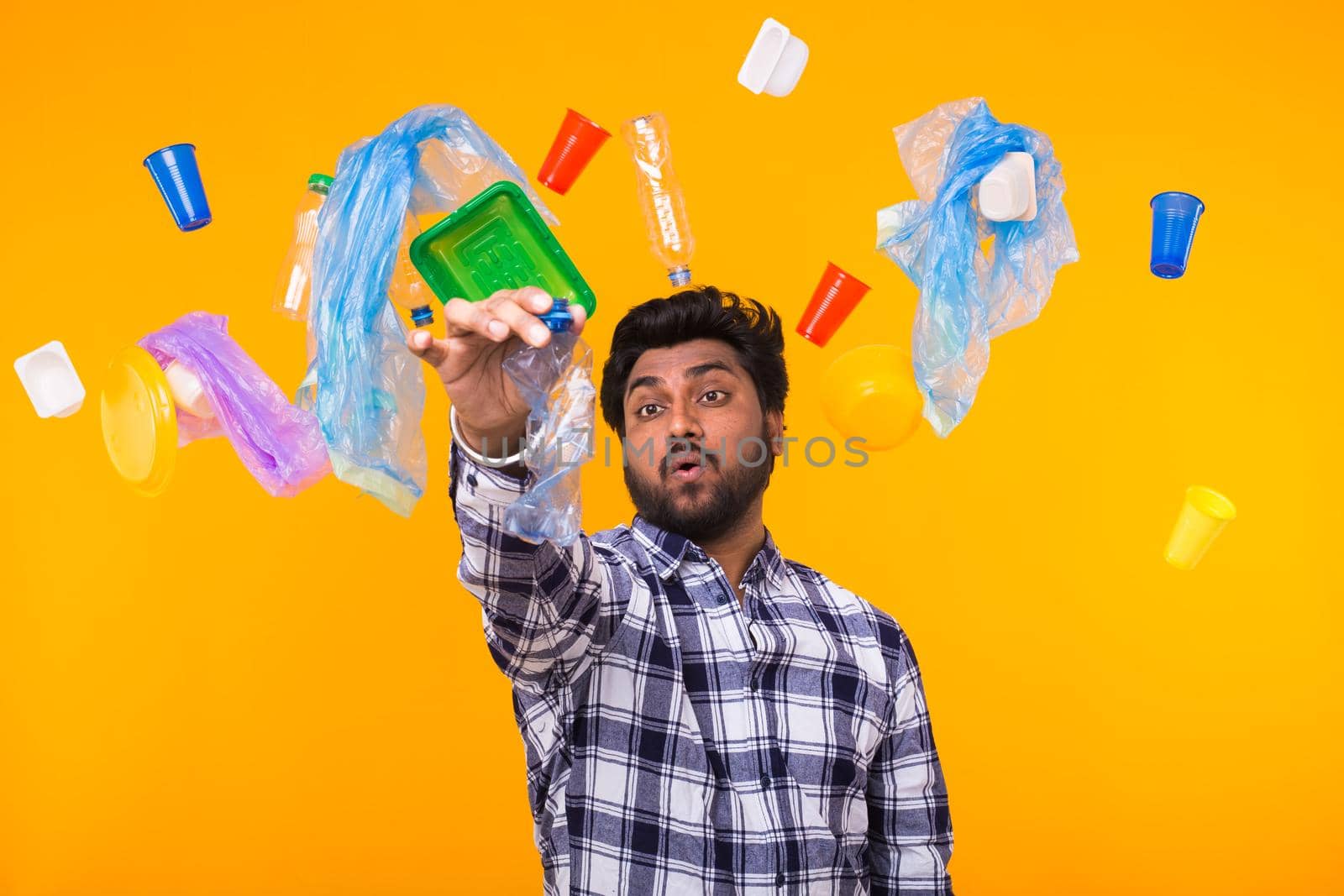 Environmental pollution, plastic recycling problem and ecology problem concept - confused Indian man holding crumpled plastic bottle on yellow background.