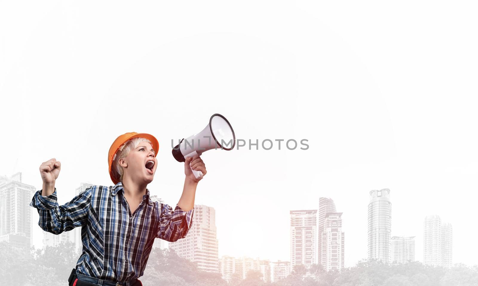 Expressive woman in safety helmet shouting into megaphone. Portrait of young emotional construction worker with wide open mouth on foggy city background. News announcement and advertisement.