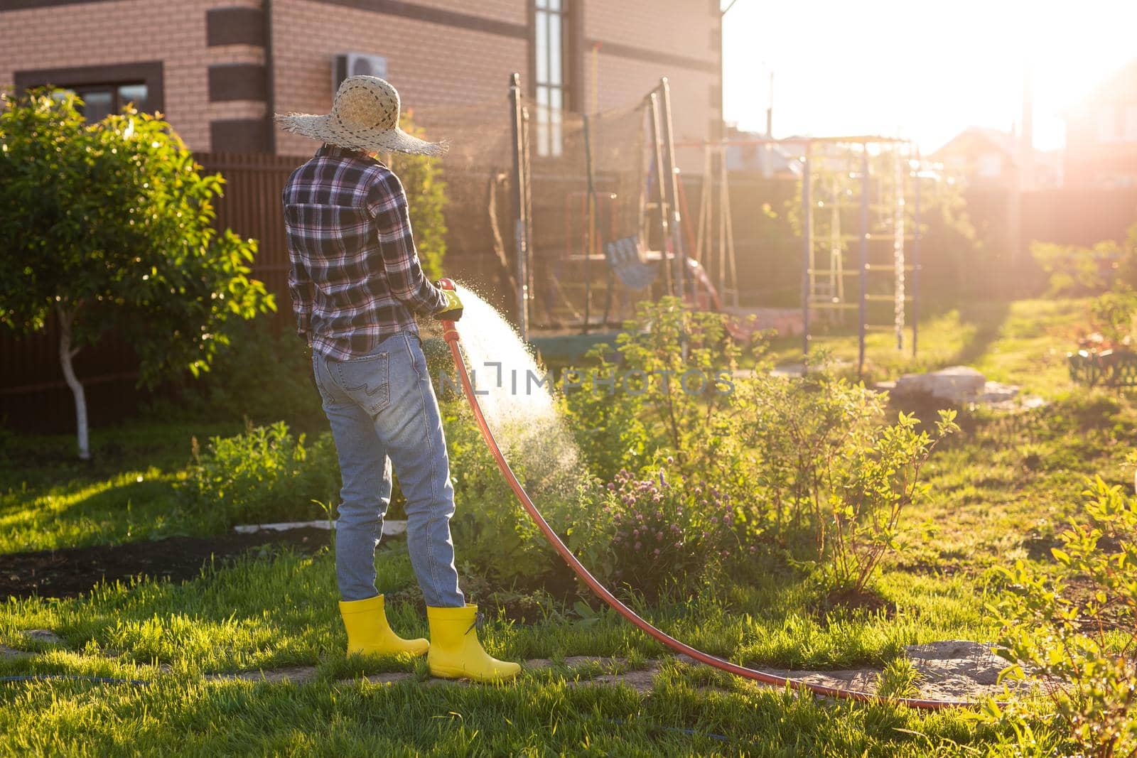 Caucasian woman gardener in work clothes watering the beds in her vegetable garden on sunny warm summer day. Concept of working in the garden