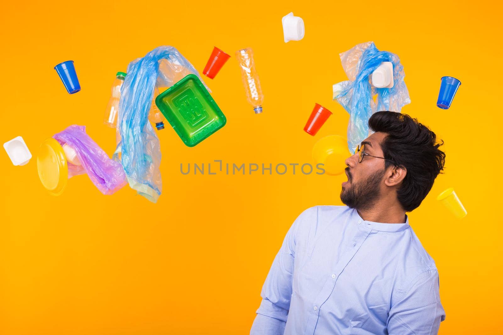 World Environment Day, plastic recycling problem and environmental disaster concept - Surprised Indian man looking on trash on yellow background.