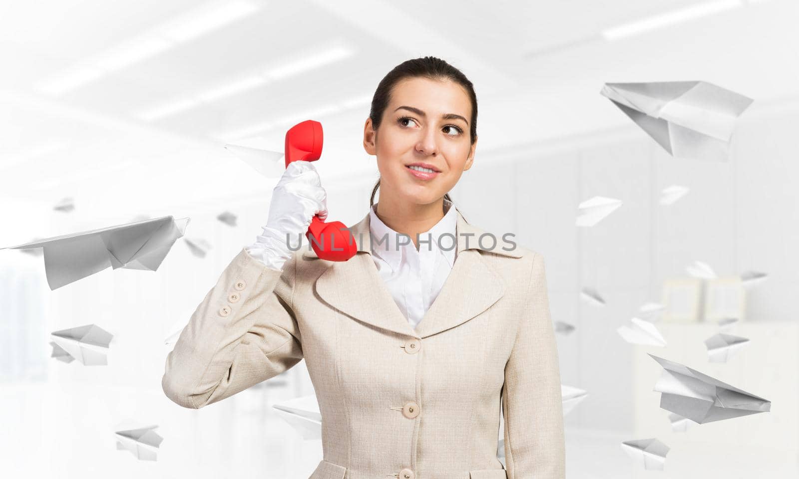 Smiling young woman holding retro red phone in office with flying paper planes. Call center operator in business suit with landline phone. Hotline telemarketing. Business assistance and consultation.