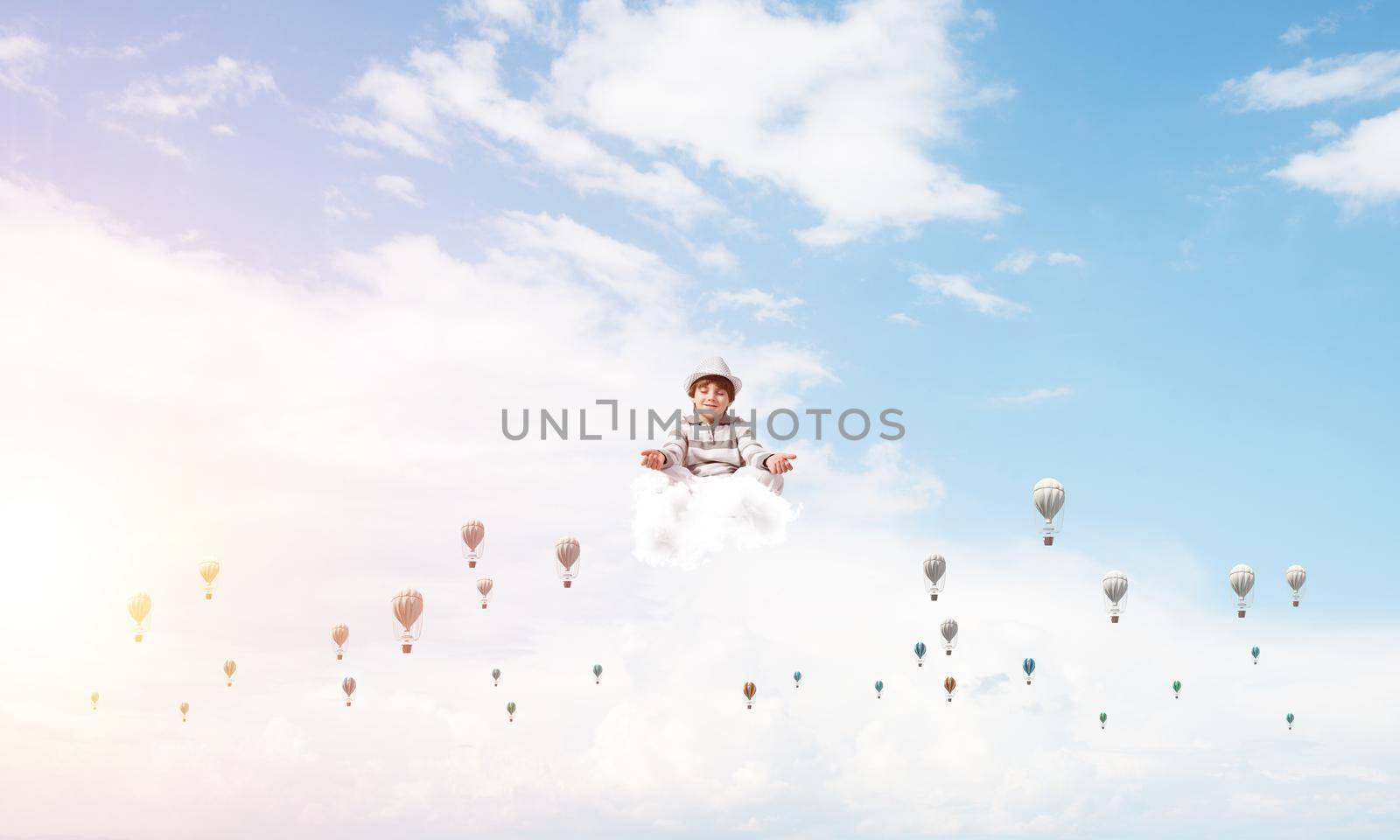 Young little boy keeping eyes closed and looking concentrated while meditating among flying balloons in the air with cloudy skyscape on background.