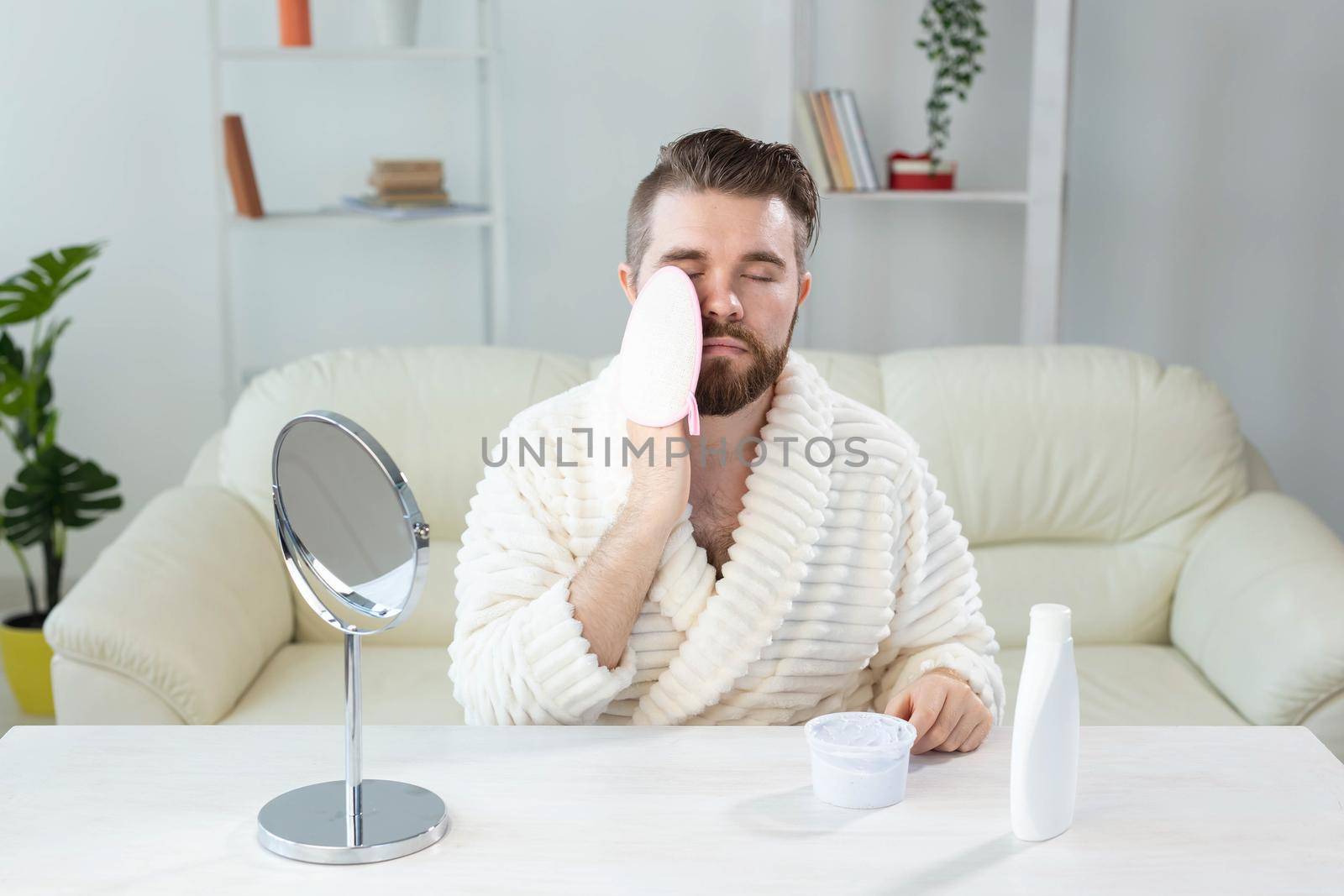 Bearded man look in mirror and clean scrub face with sponge, happy millennial male metrosexual do beauty procedures at home, take care of healthy skin, skincare concept.
