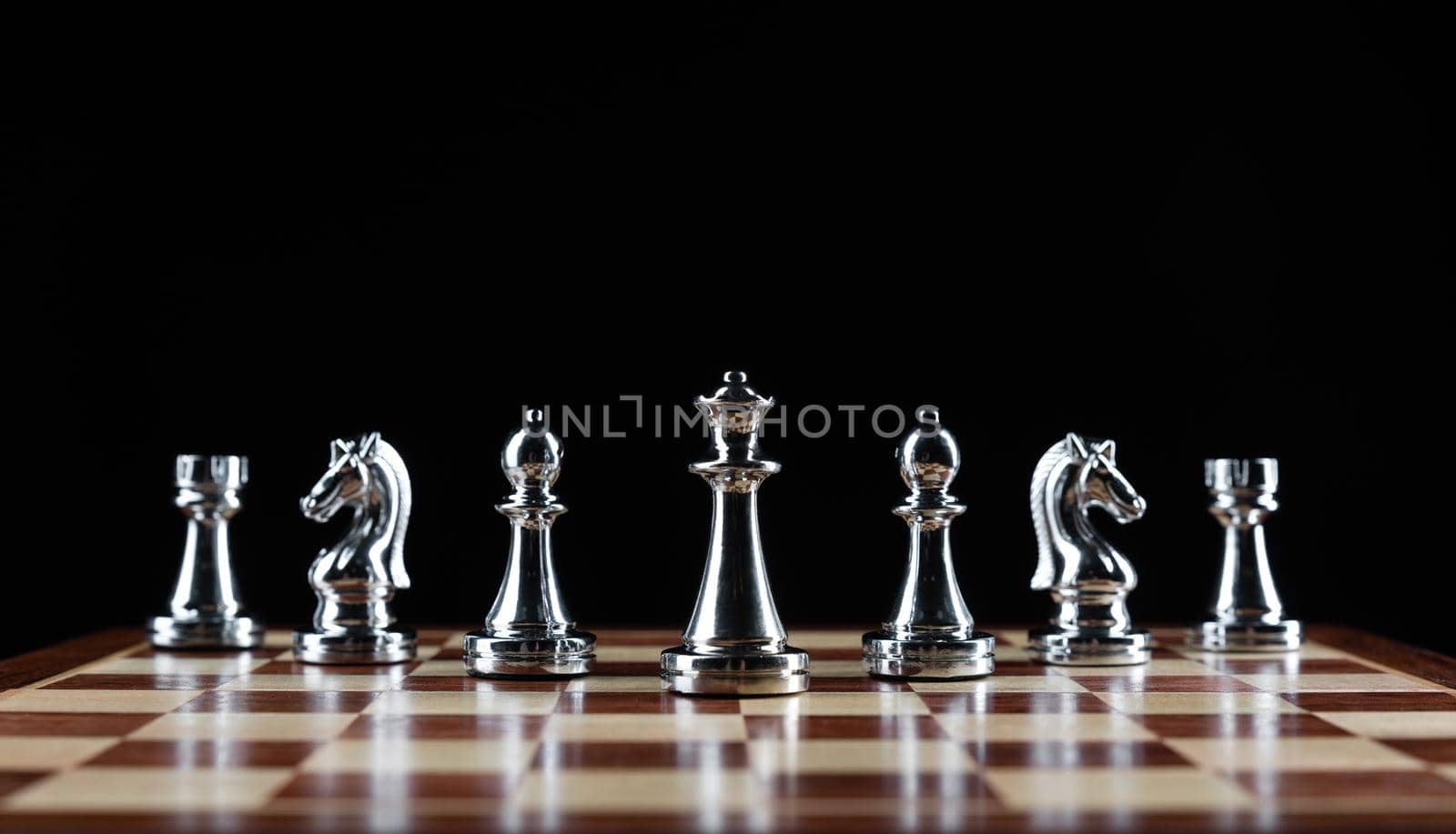 Shiny steel chess figures standing on wooden chessboard. Intellectual duel and tactical battle symbol. Strategy planning and corporate leadership concept. Silver metal chess pieces on black background