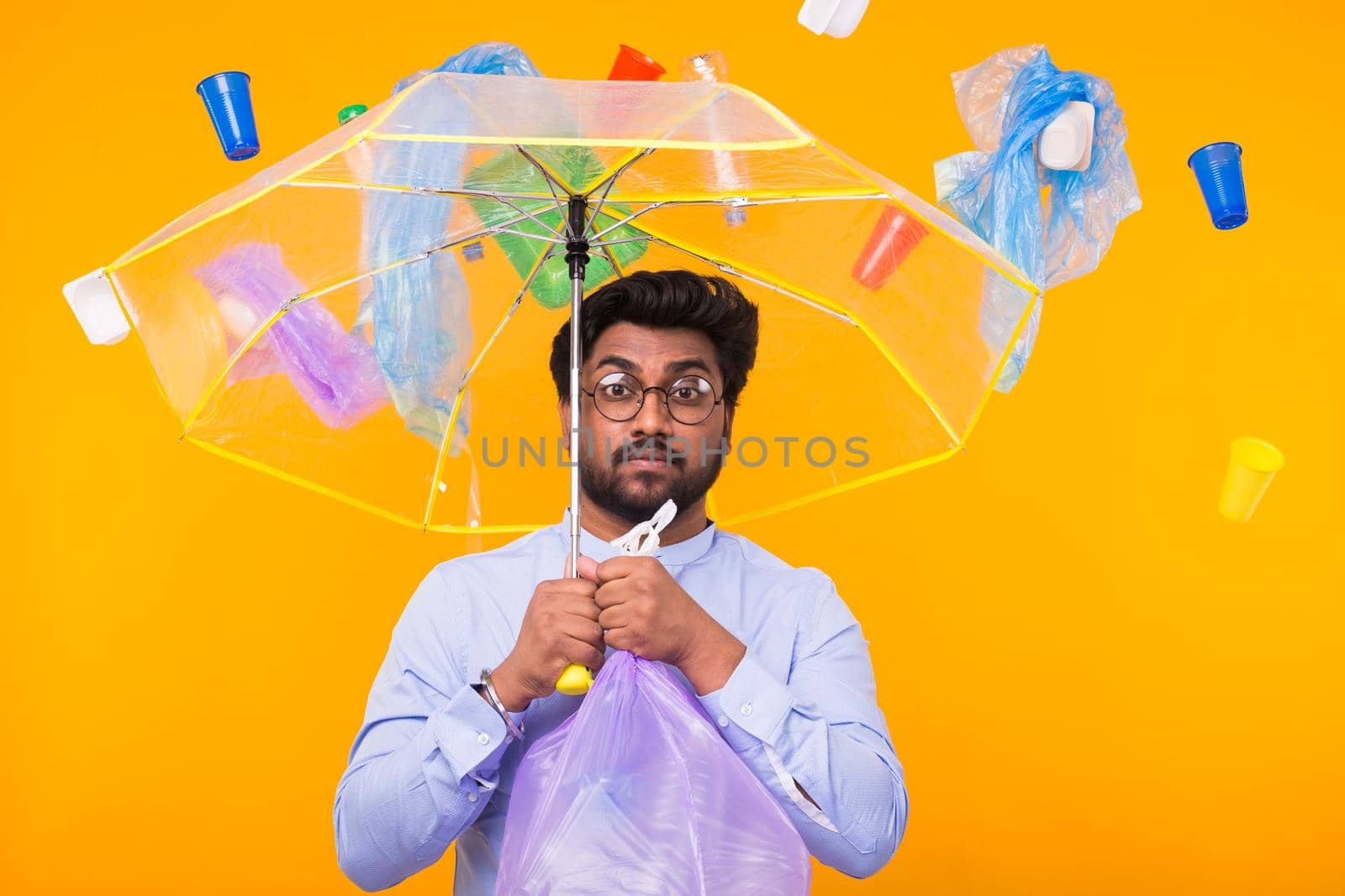 World Environment Day, plastic recycling problem and environmental disaster concept - Sad Indian man hiding from garbage under an umbrella on yellow background.