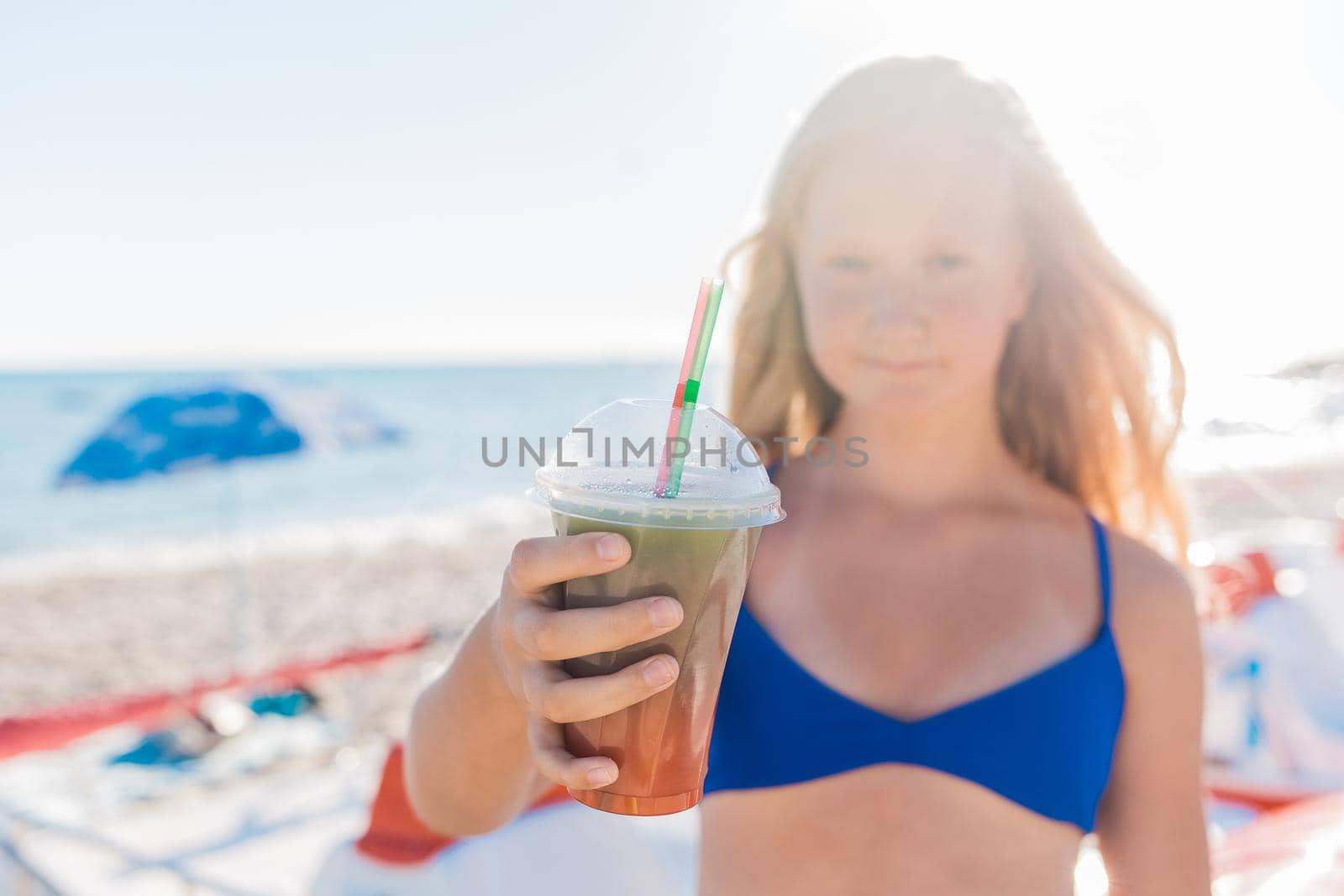 A young girl with blonde hair of European appearance, a teenager holds a colored cold non-alcoholic cocktail in her hand against the background of the sea beach.