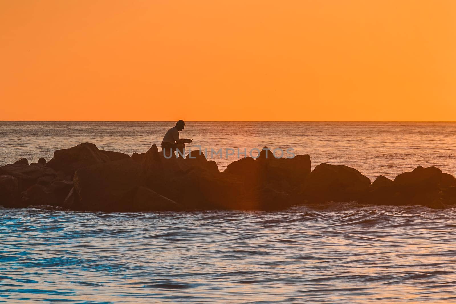 The silhouette of a lonely man sitting in meditation on the rocks of a breakwater near the sea coast against the orange sunset.