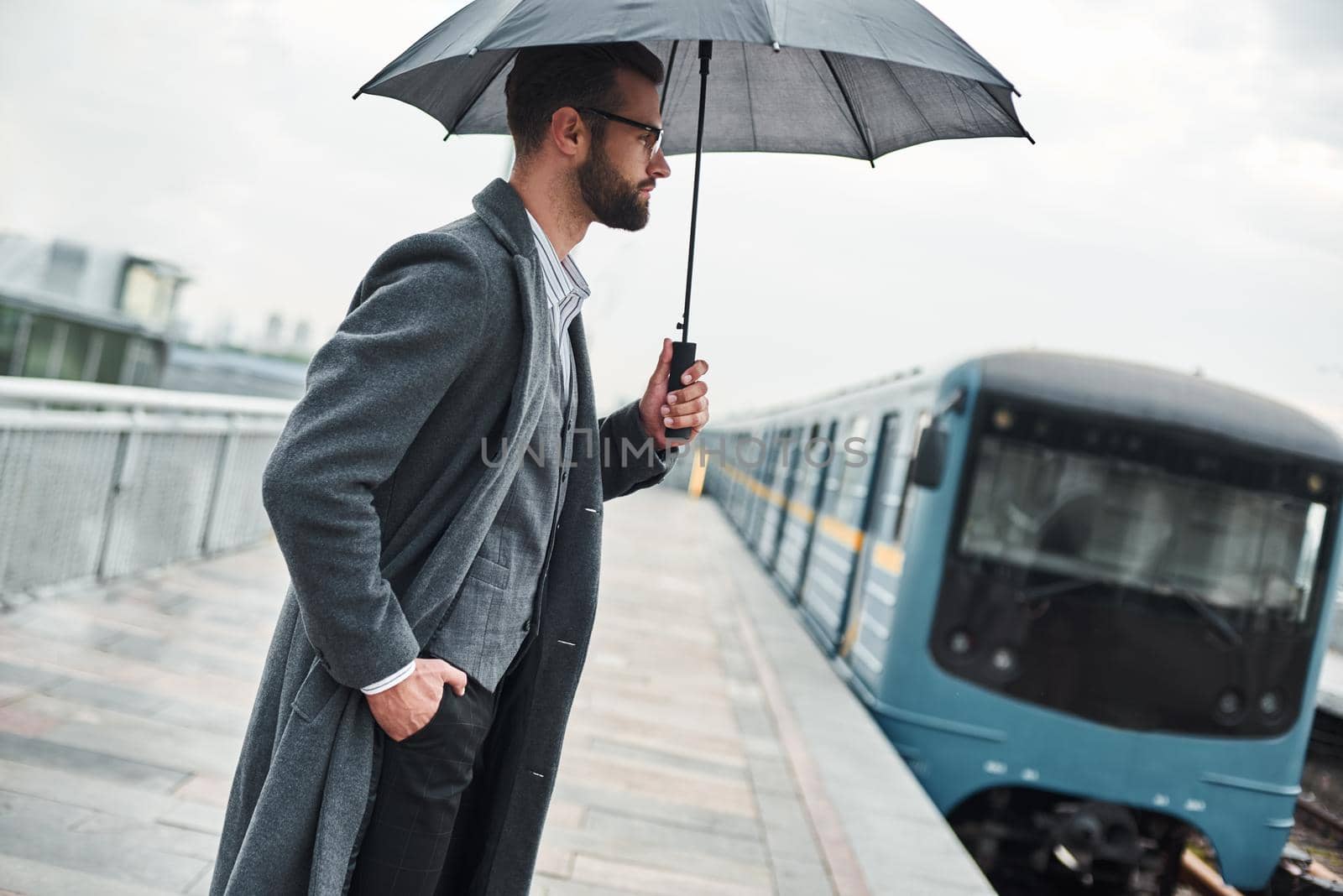 Waiting for train. Young businessman standing near railway under umbrella