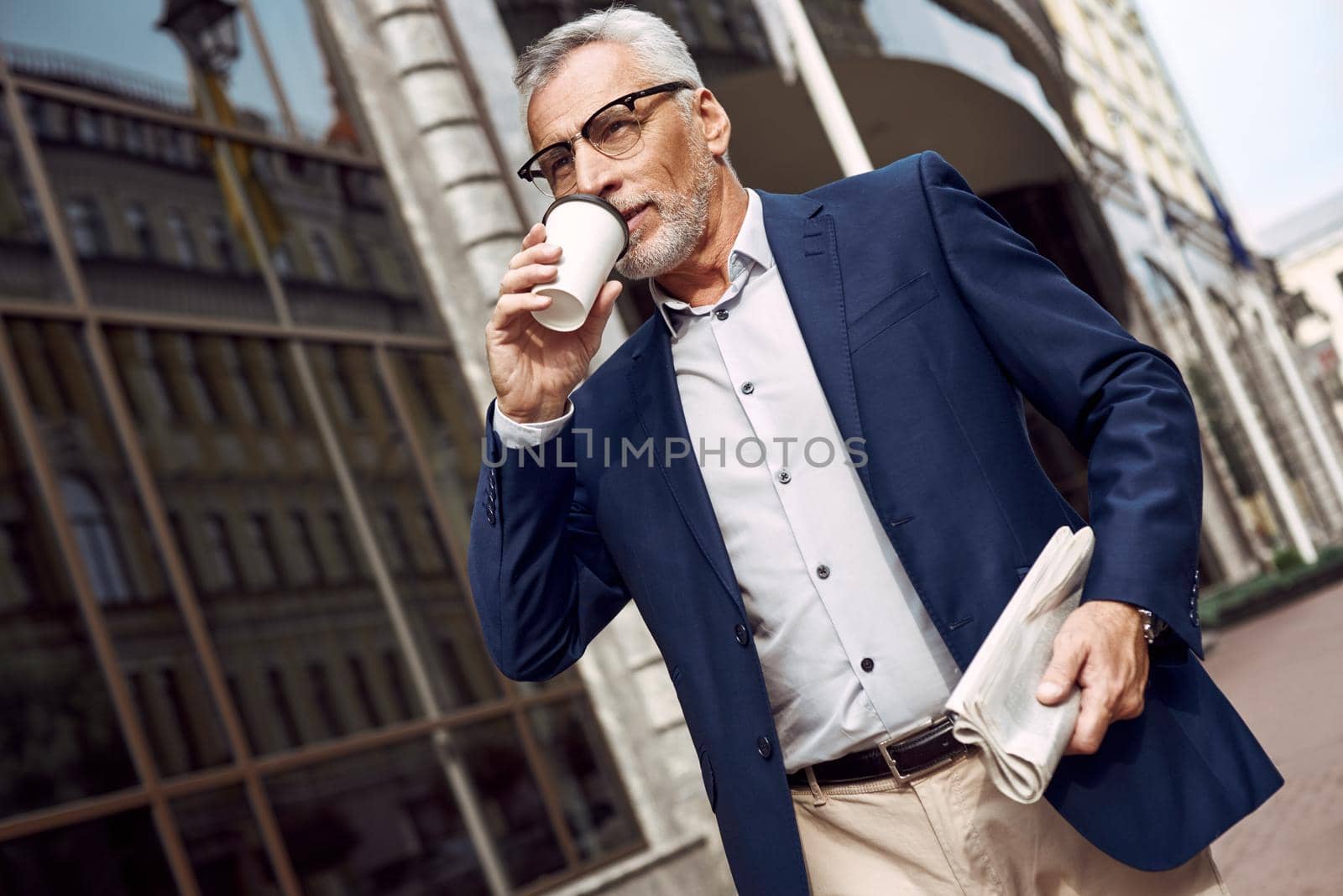 Handsome senior man in smart casual wear carrying disposable cup while walking through the city street