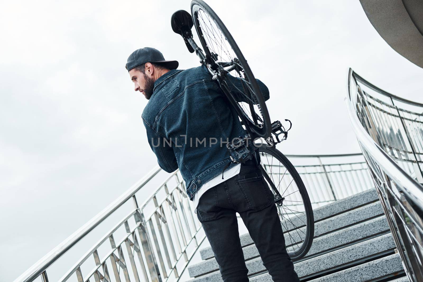 Outdoors leisure. Young stylish man walking up the stairs holding bicycle looking aside curious