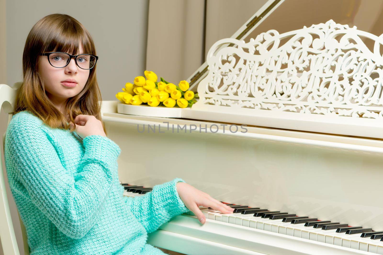 A sweet schoolgirl girl sits beside a white grand piano on which lies a bouquet of flowers. The concept of studying music, spring mood, happy people.