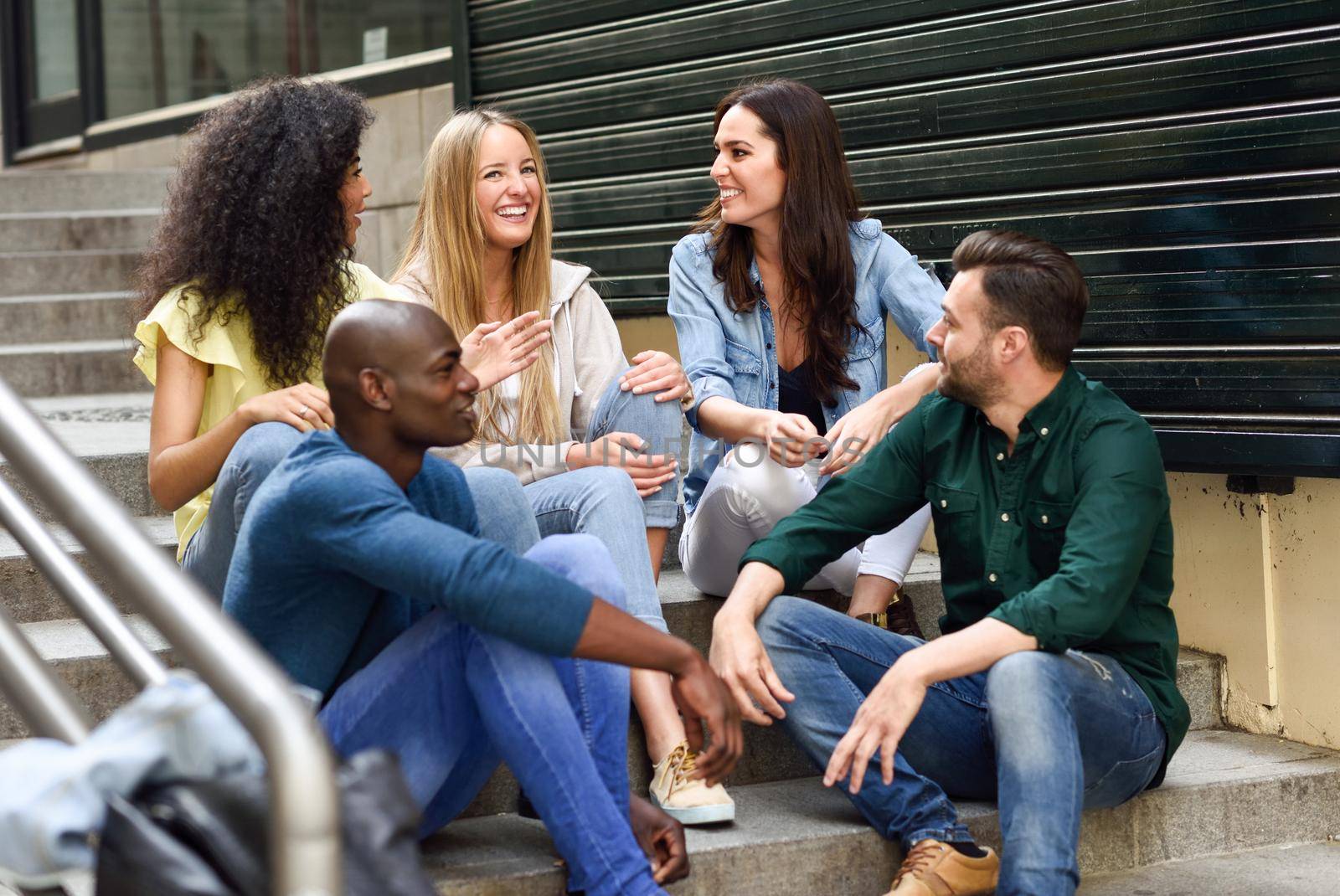 Multi-ethnic group of young people having fun together outdoors in urban background. group of men and woman sitting together on steps.