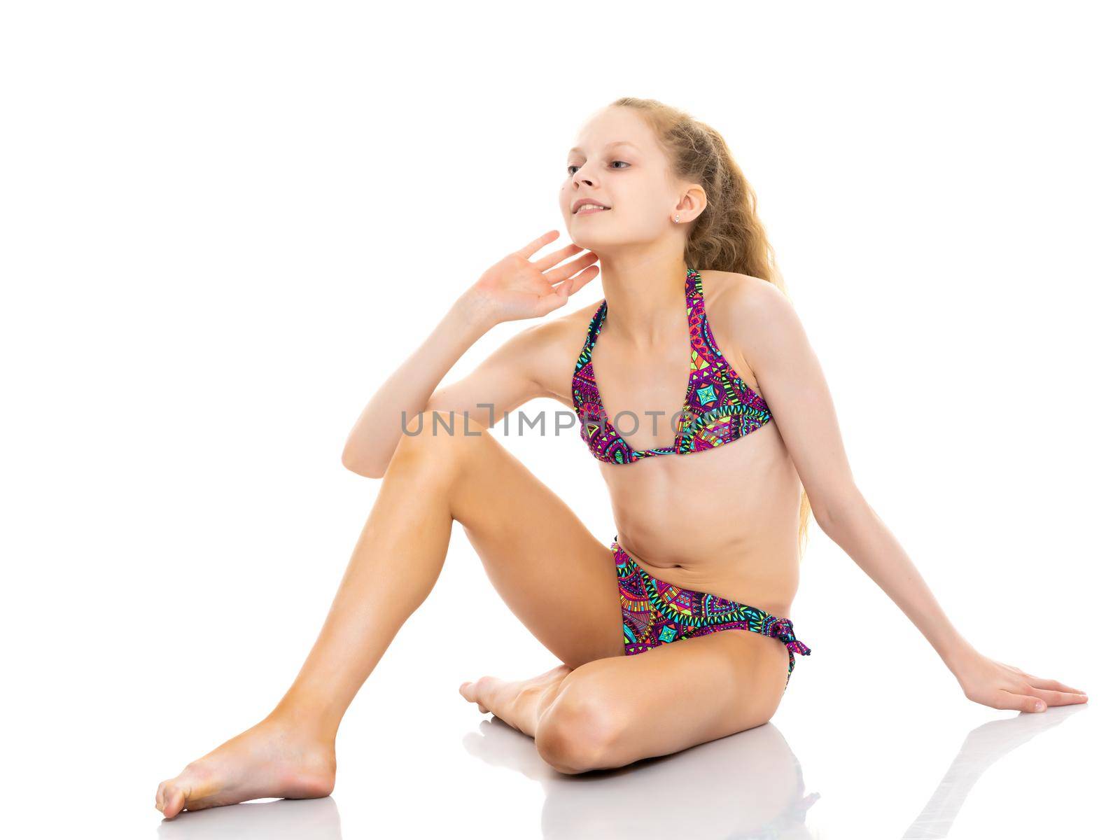 A beautiful little school girl in a bathing suit is sitting on the studio floor on a white background. The concept is suitable for illustrating summer children's and family recreation at sea, sun tanning on the beach.