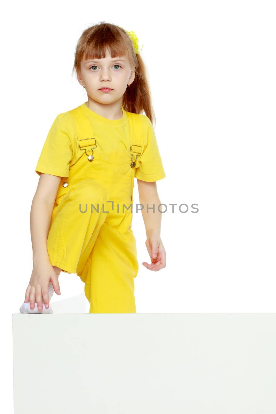 Girl with a short bangs on her head and bright yellow overalls.She put her foot on the white advertising banner.