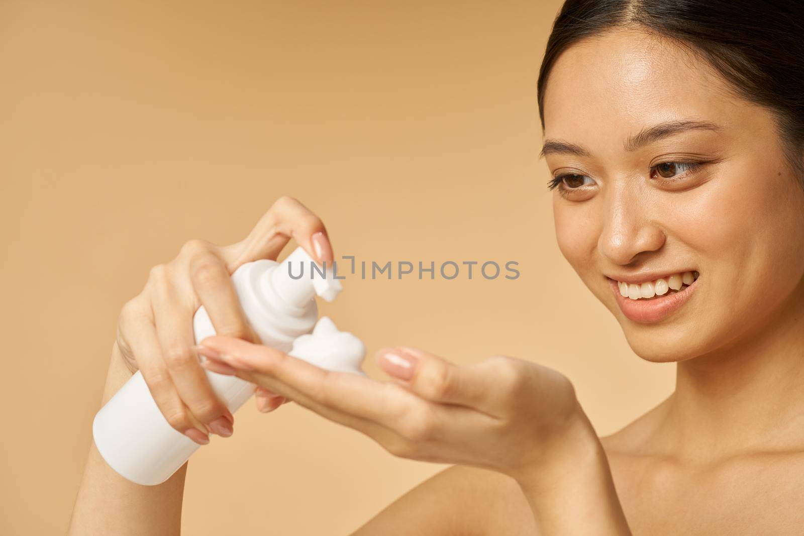 Close up portrait of cute young woman smiling, holding a bottle of gentle foam facial cleanser isolated over beige background. Beauty products and skin care concept