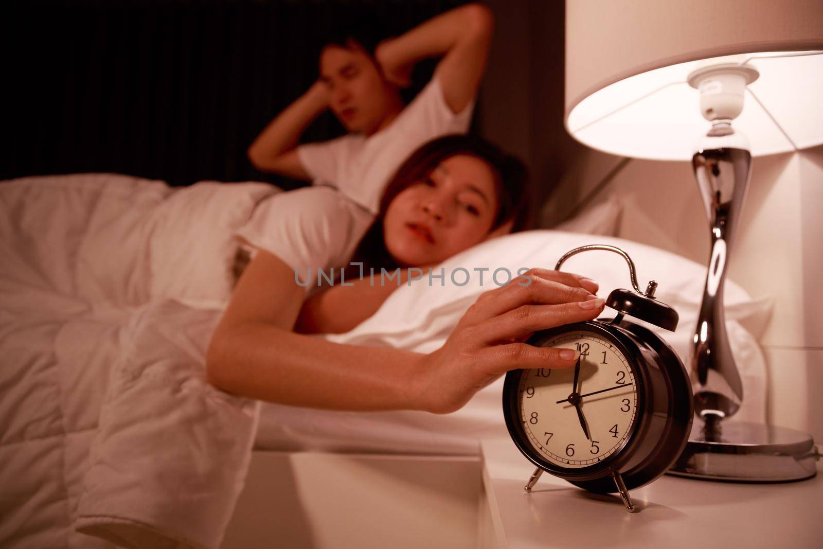 sleepy young couple in bed with extending hand to alarm clock in the morning