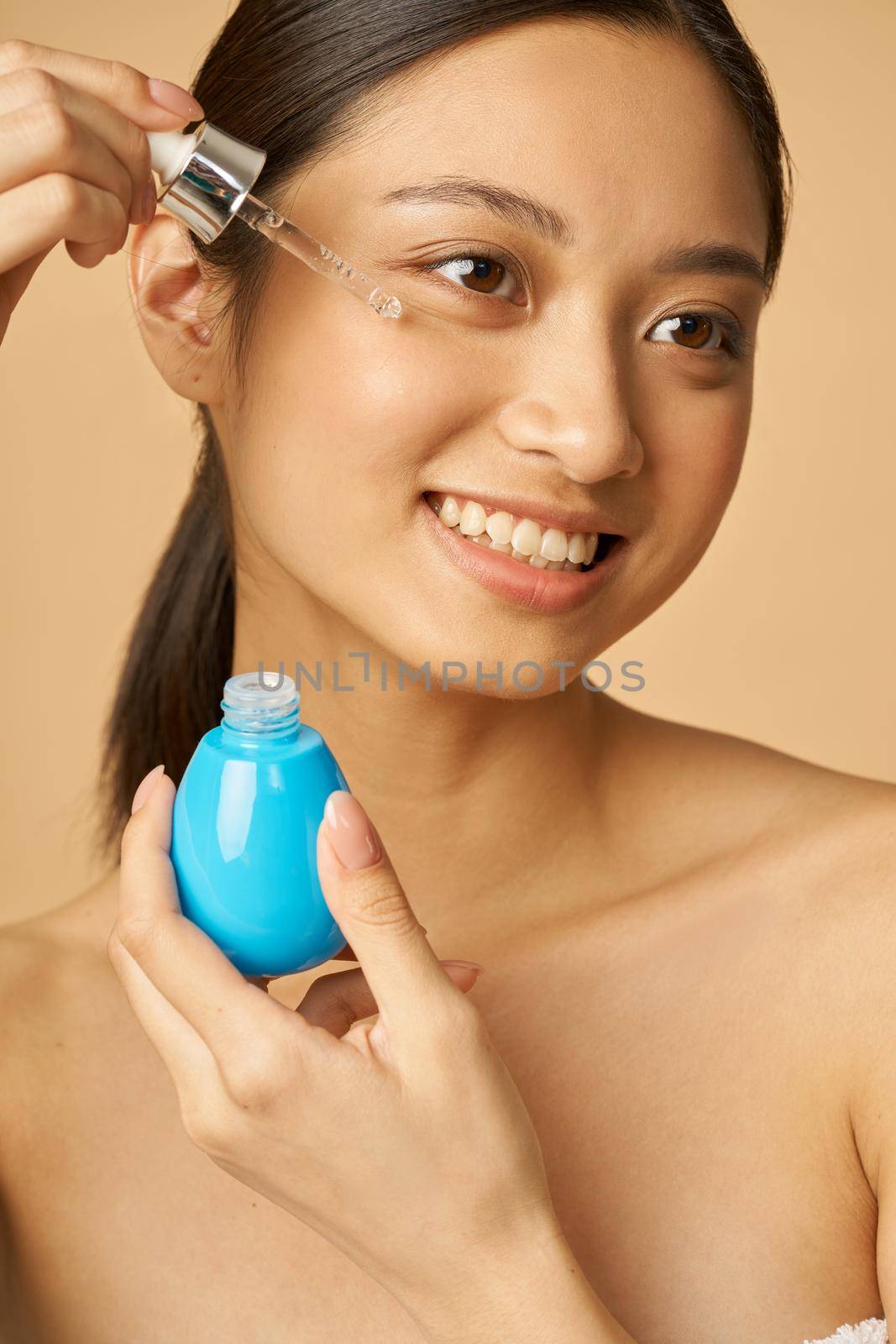 Portrait of cheerful young woman dropping oil serum with pipette on face for fresh perfect skin, posing isolated over beige background by friendsstock