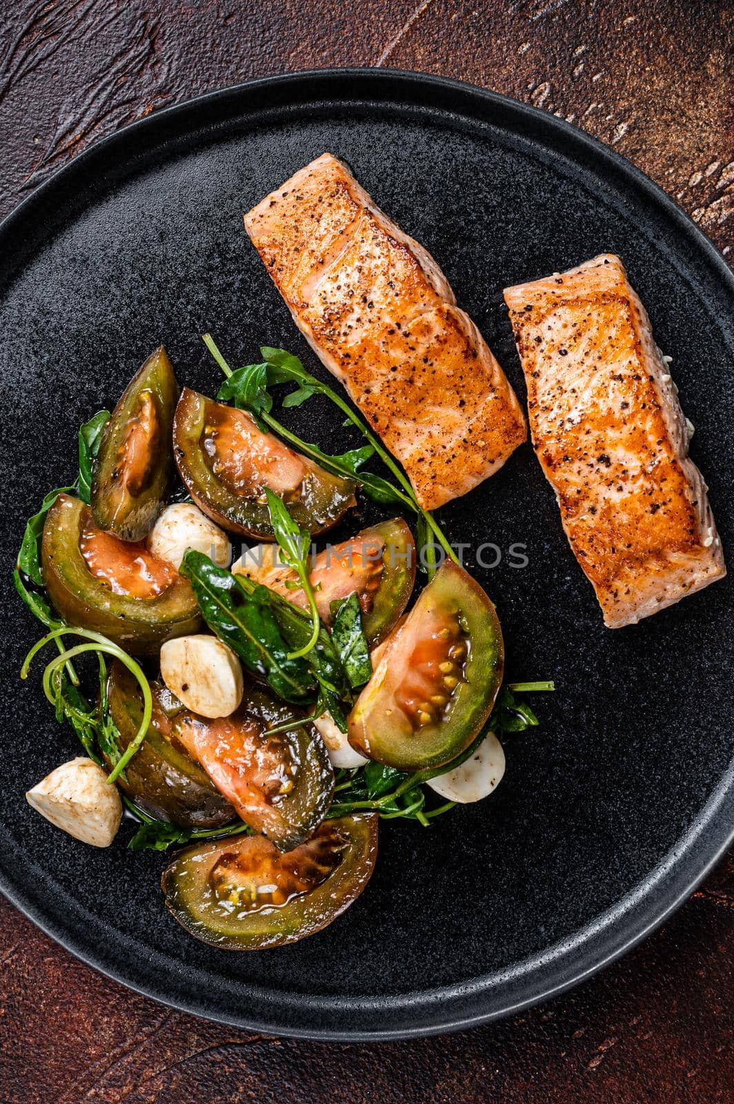 Grilled Salmon Fillet Steaks with arugula and tomato salad on a plate. Dark background. Top view.