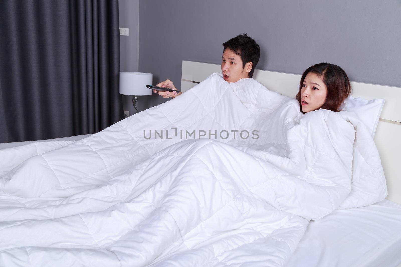 young couple watching scared movie with tv remote control under the blanket on bed