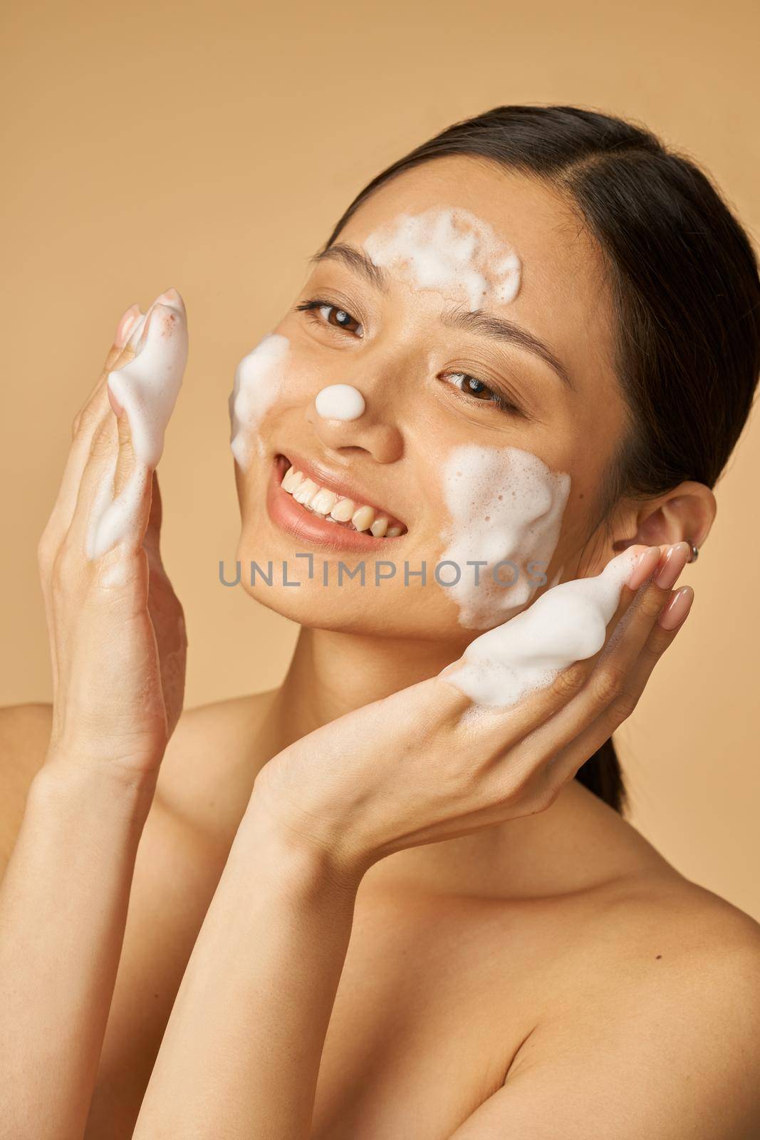 Beauty portrait of joyful young woman smiling while applying gentle foam facial cleanser isolated over beige background by friendsstock