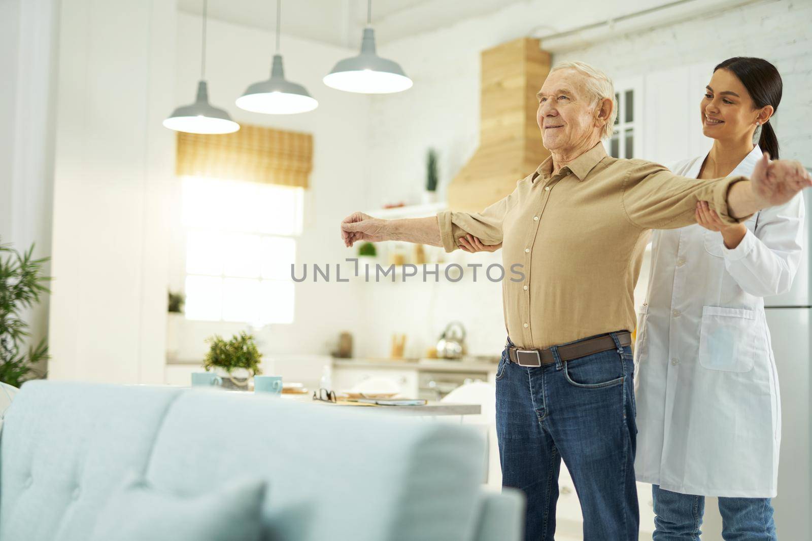 Happy female doctor in white coat helping an elderly man to do exercises in his apartment